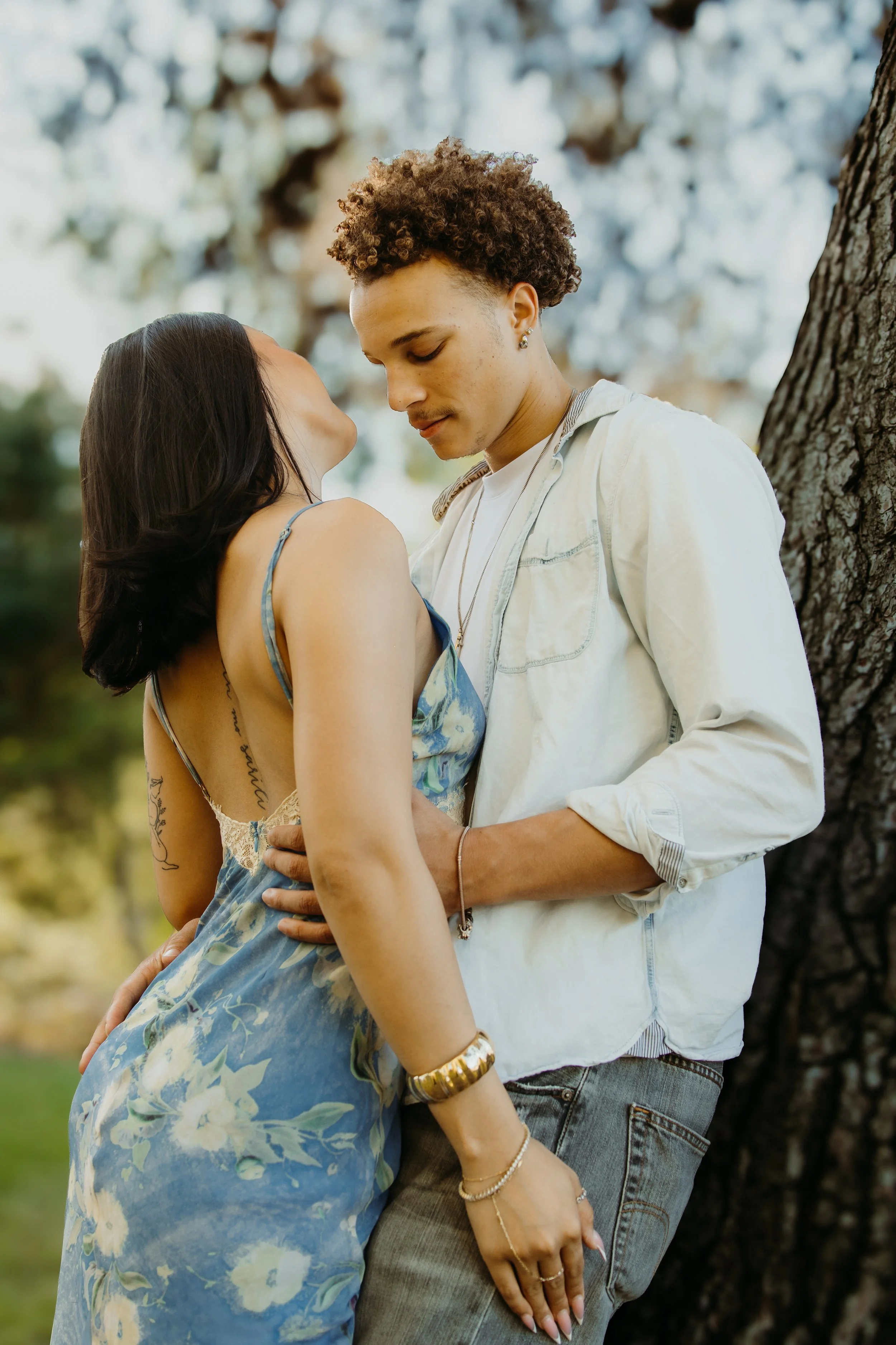 A young couple standing close together outdoors near a tree, with their foreheads touching and eyes closed, appearing to be in a romantic moment.