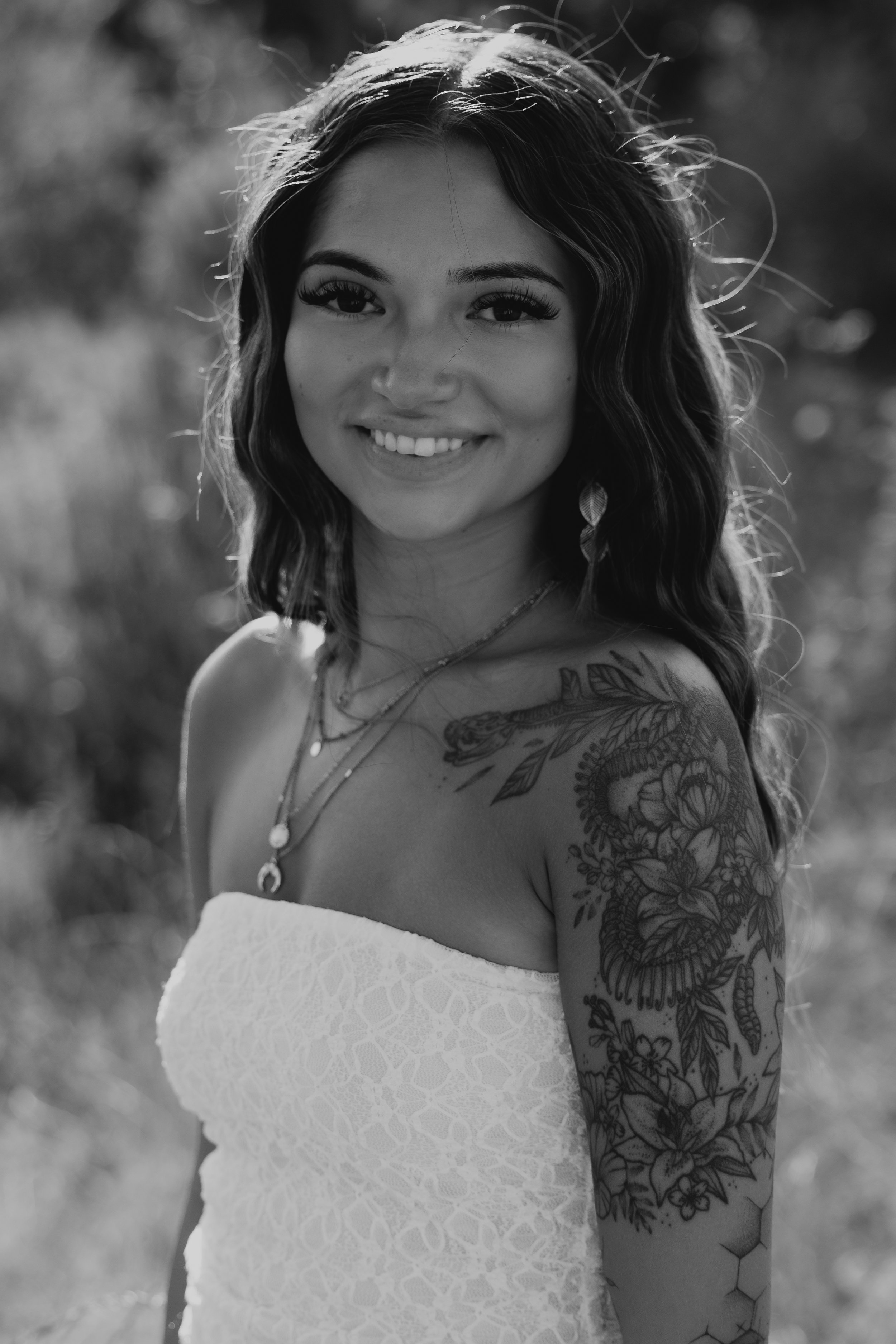 Black and white photo of a smiling woman with wavy hair, wearing a strapless dress and displaying tattoos on her left arm and shoulder.