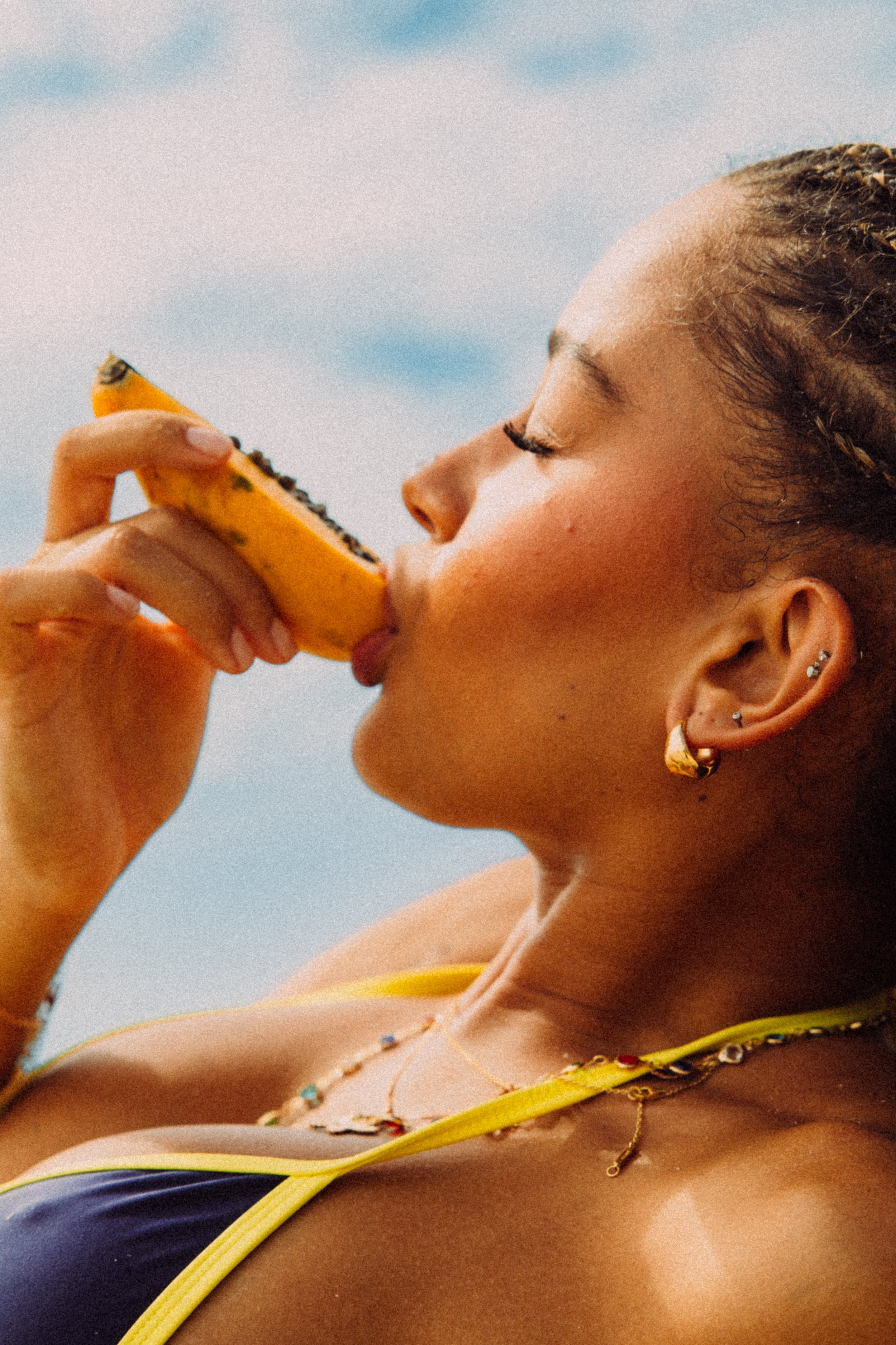A woman with braided hair, wearing earrings and a necklace, is holding a slice of banana with chocolate chips close to her face, with her eyes closed, against a bright outdoor sky background.