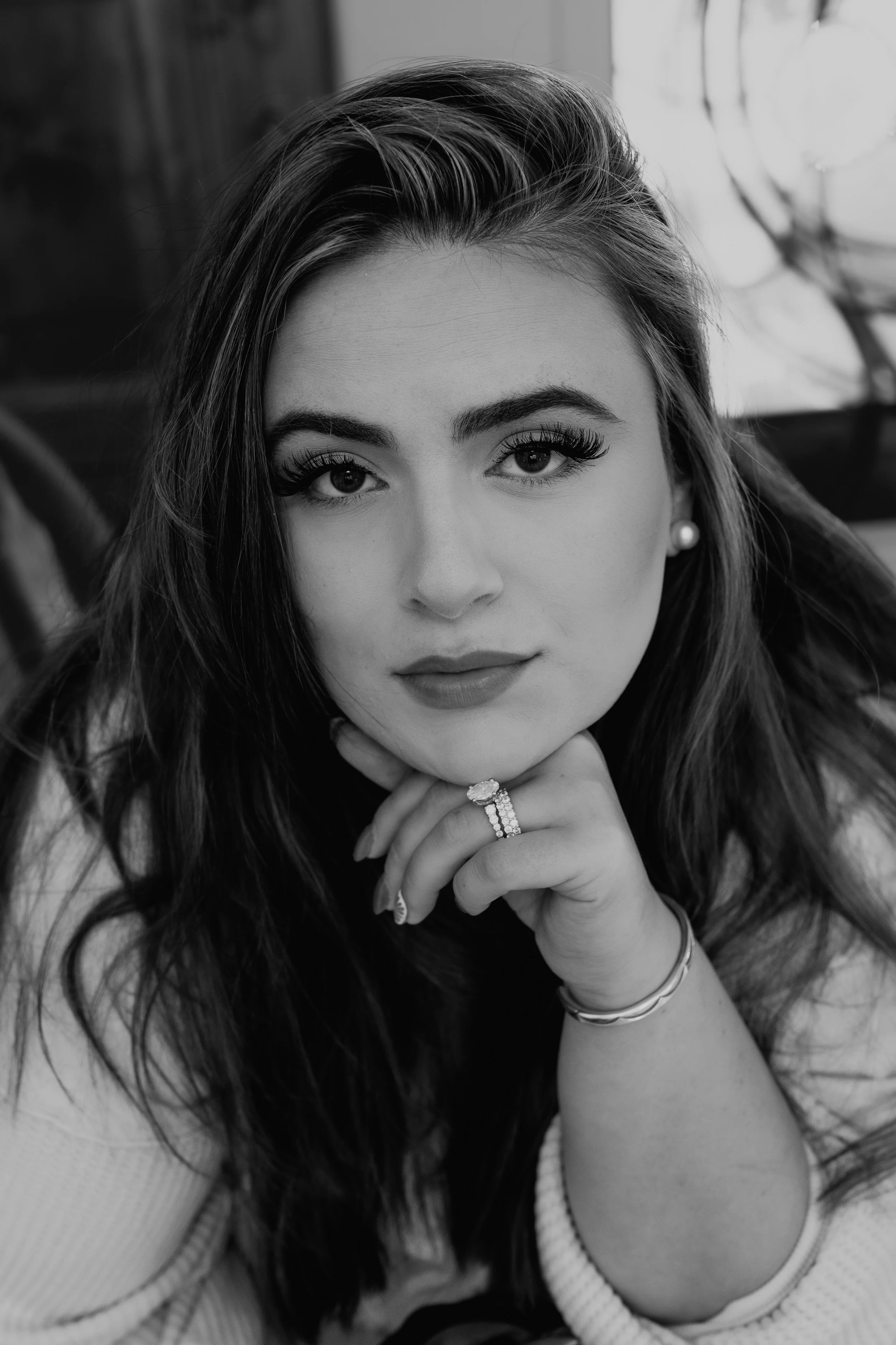 Close-up black and white photograph of a woman with long hair, wearing makeup, earrings, and rings, resting her chin on her hand, with a blurred background.