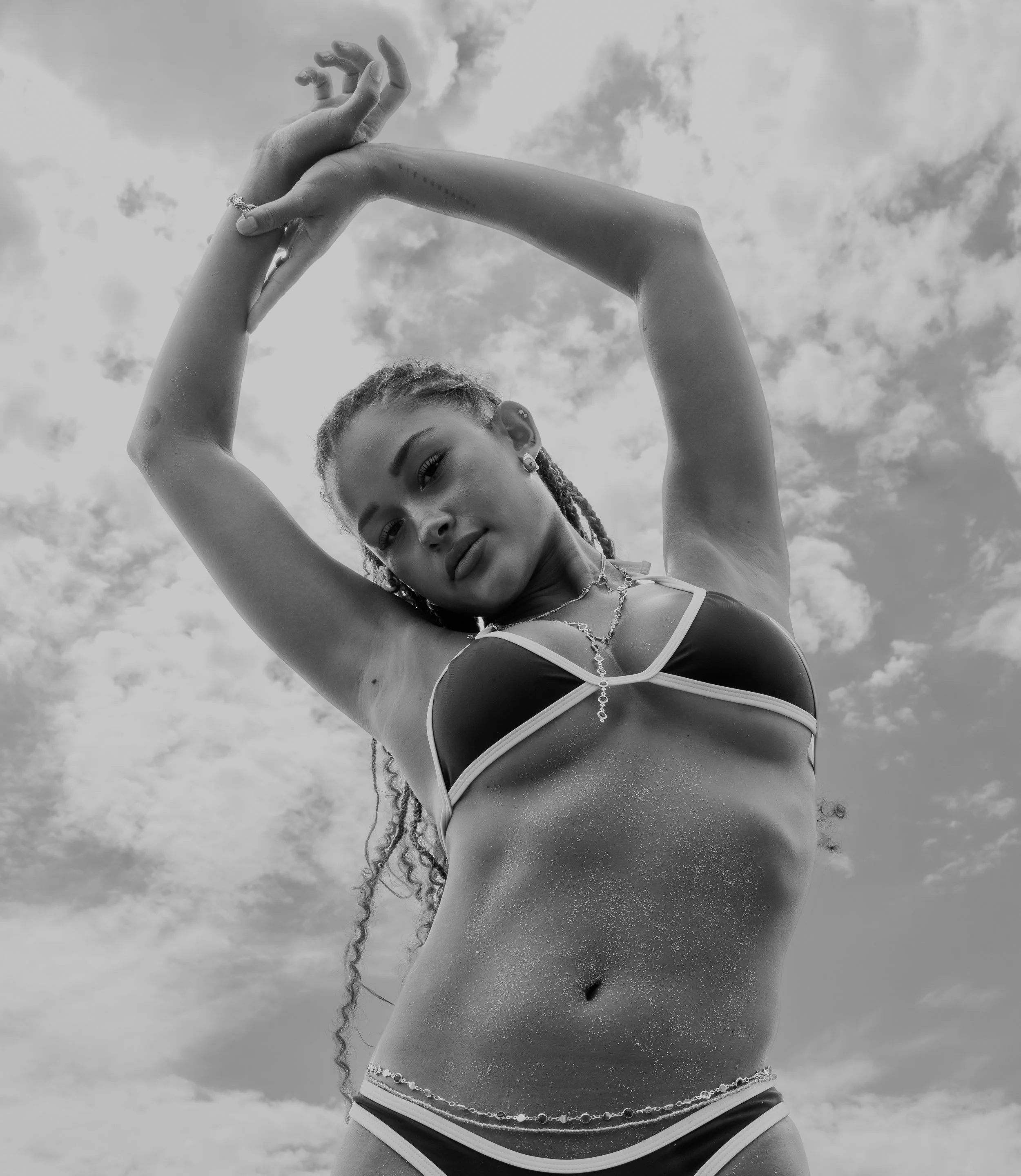 Black and white photo of a woman with braided hair posing outdoors with arms raised and hands clasped above her head, looking towards the camera with clouds in the sky background.