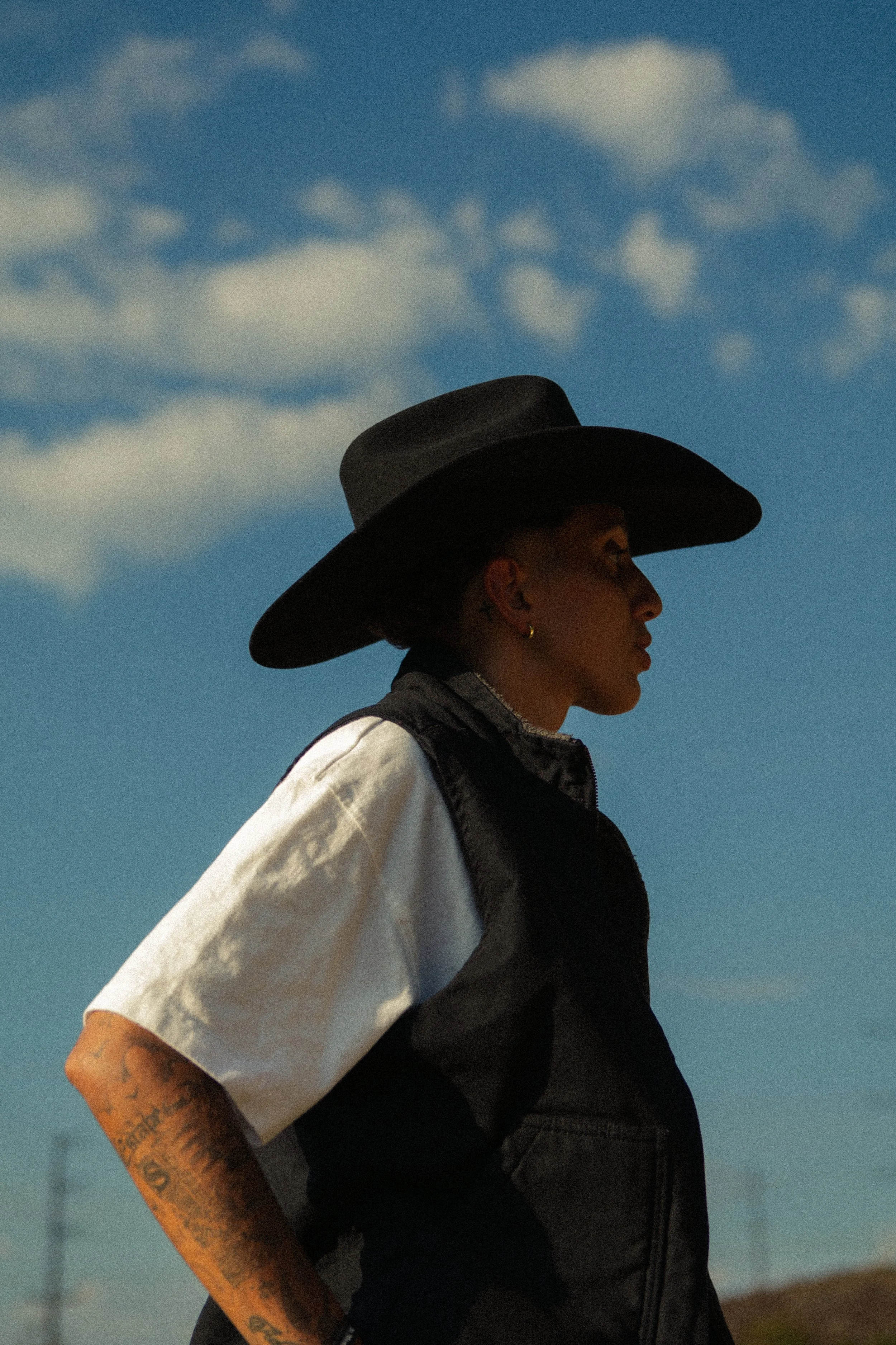 A person wearing a large black hat, a black vest over a white and gray shirt, with tattoos on their left arm, standing against a bright blue sky with scattered clouds.