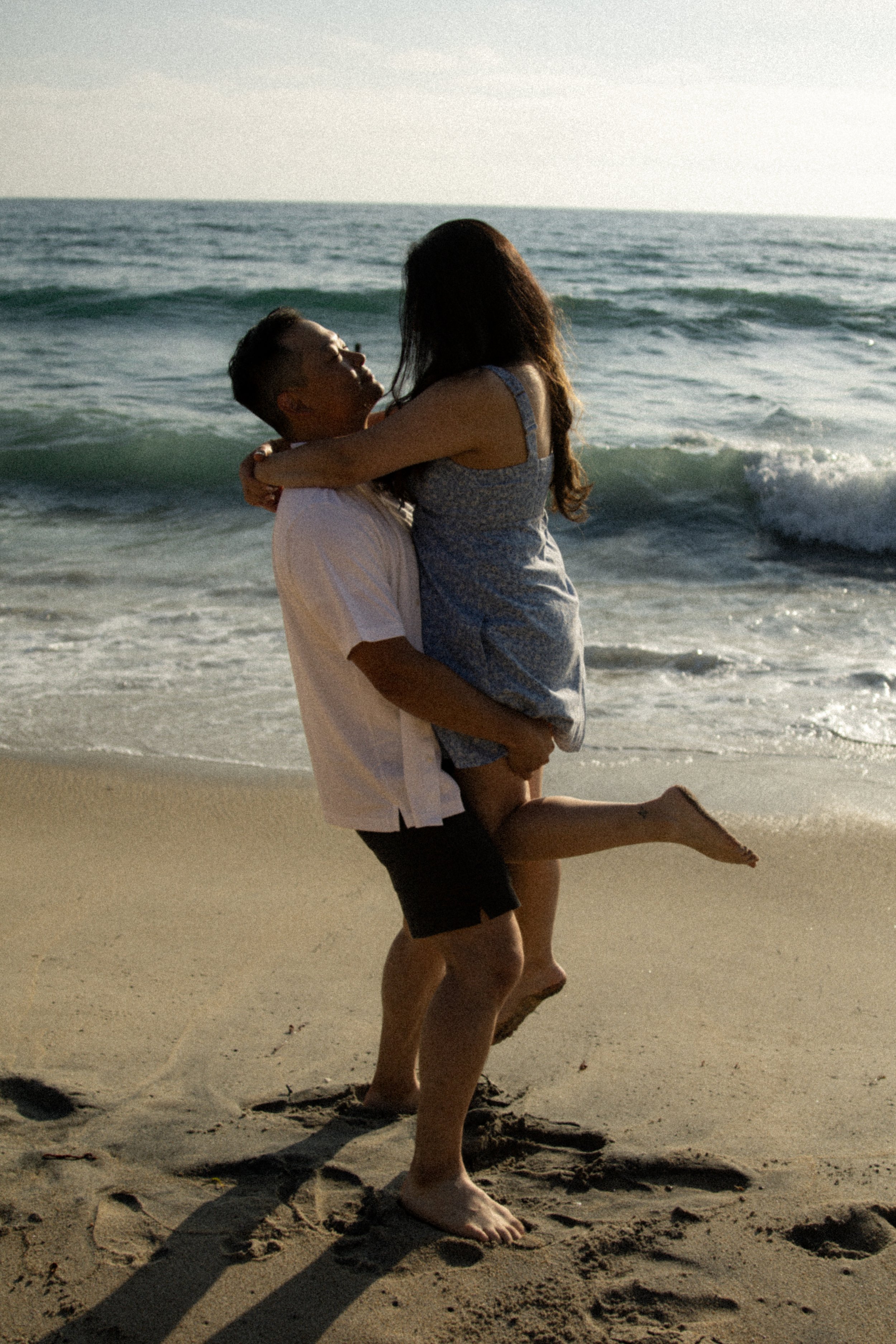 A couple at the beach during sunset, with the man lifting the woman in his arms as they smile at each other. The waves are in the background.