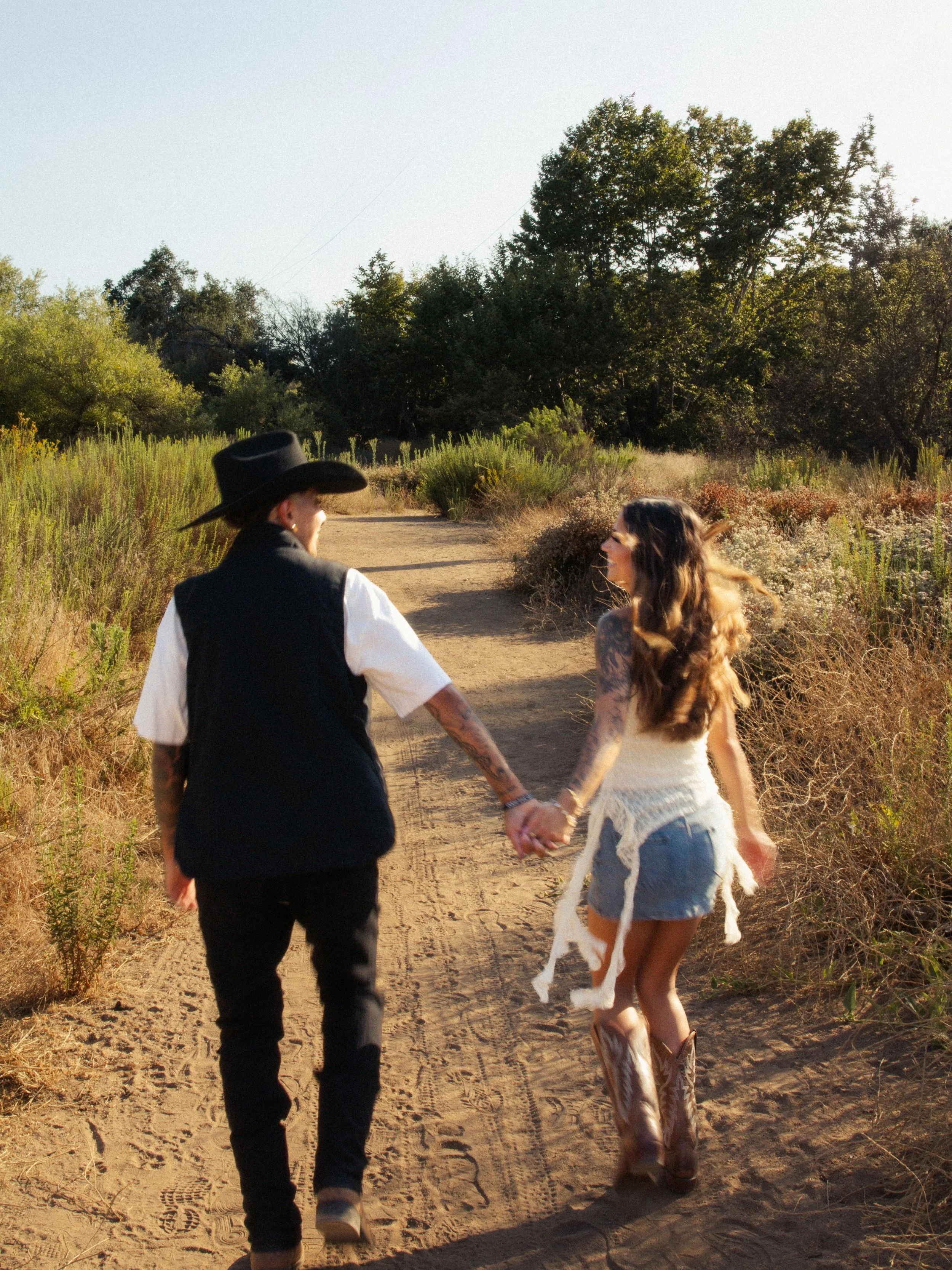 A man wearing a black cowboy hat and black vest walking hand-in-hand with a woman in a white tank top and denim shorts on a dirt trail surrounded by greenery and trees.