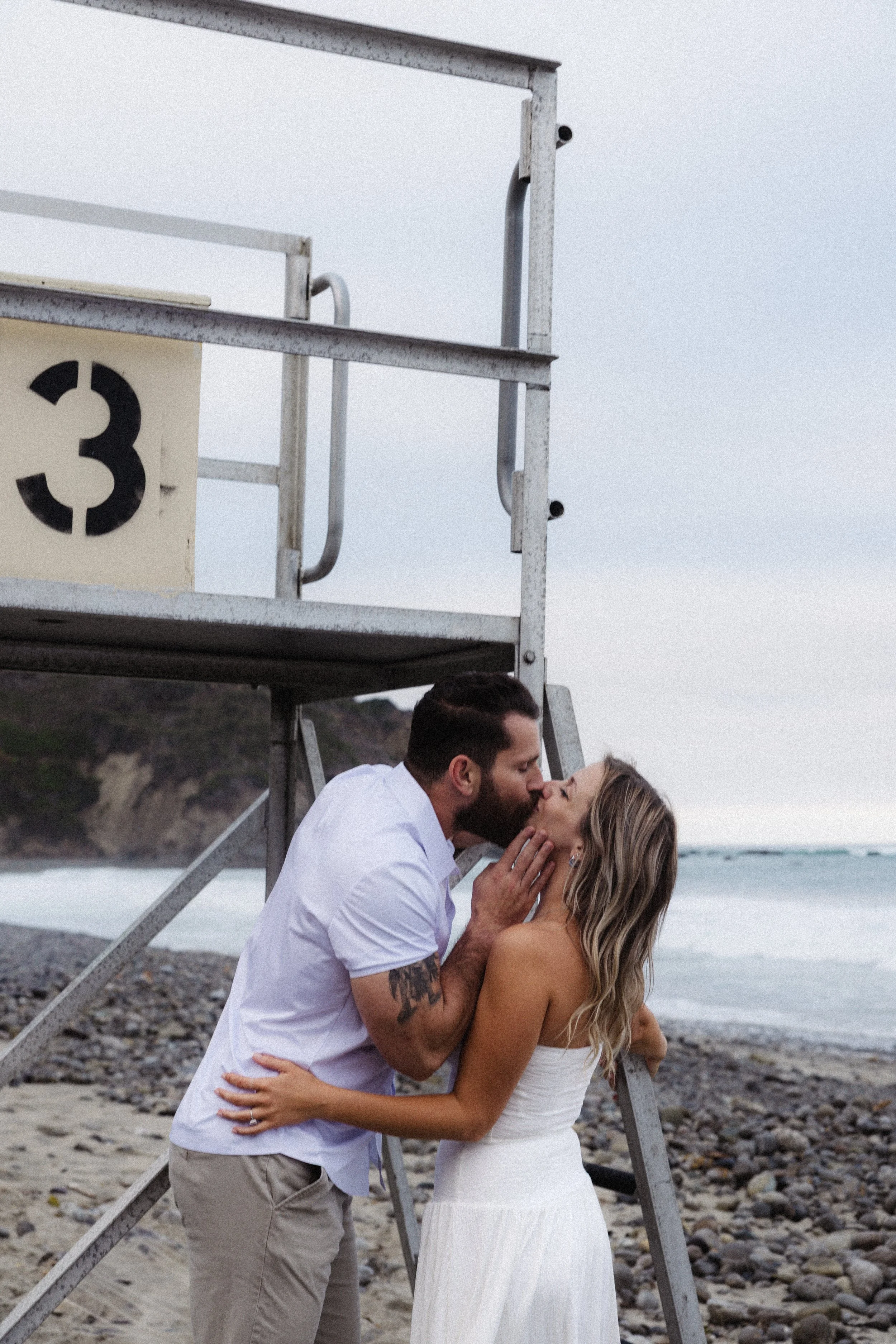 A couple sharing a kiss on a beach near a lifeguard station. The man is wearing a white shirt and beige pants, and the woman is in a white strapless dress. The background shows a rocky shoreline and the ocean, under a cloudy sky.