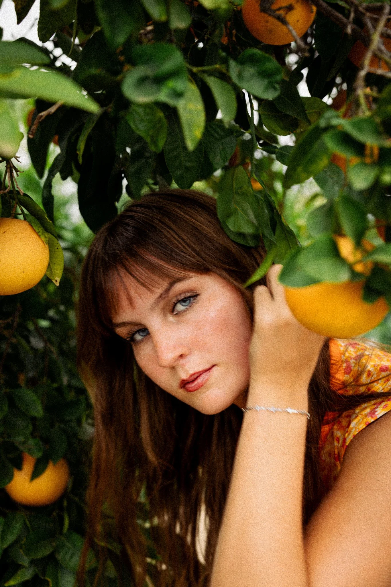 A woman with long brown hair and blue eyes is partially visible, posing among green leaves and yellow fruit on a tree, with one eye closed and her head resting on her hand.