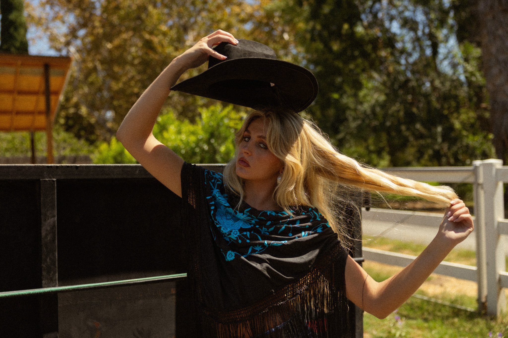 A woman with long blonde hair, wearing a black fringed dress and blue patterned scarf, raises a black hat above her head while leaning on a black fence outdoors during daytime. She looks at the camera with a slight pout, with trees and a playground i