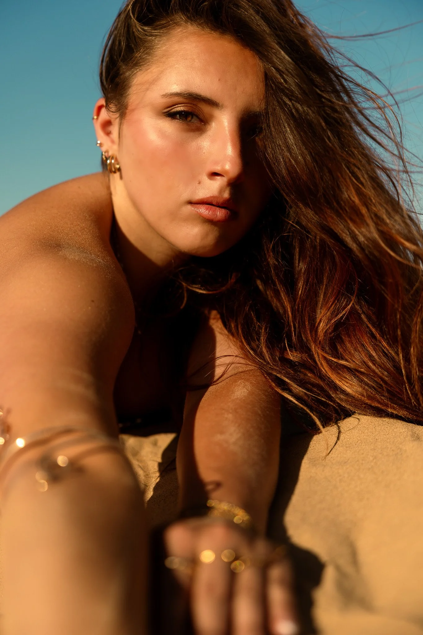 A close-up shot of a woman with long, wavy brown hair and earrings, taking a selfie outdoors with a blue sky background, illuminated by warm sunlight.