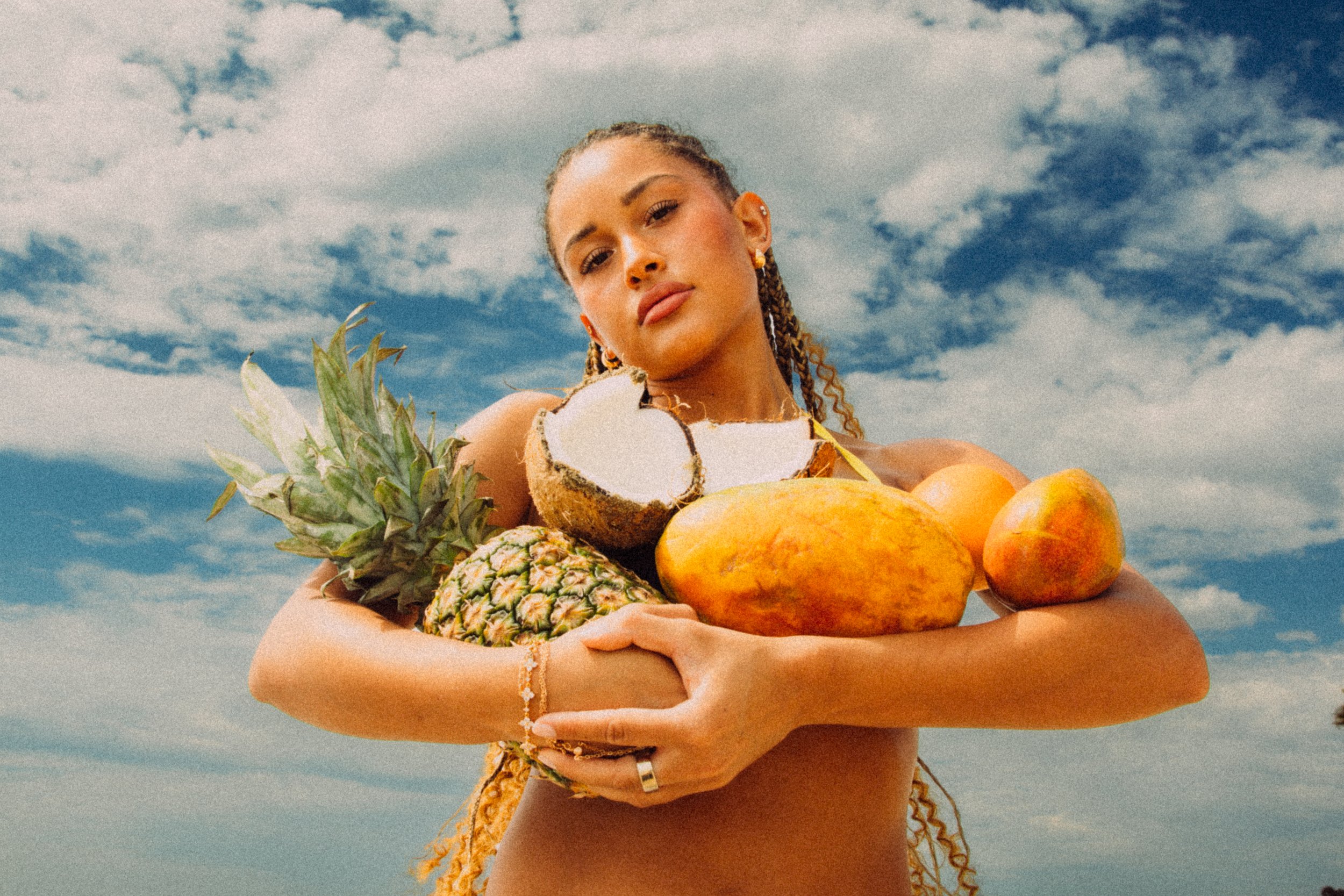 A woman holding a large assortment of tropical fruits, including pineapple, coconuts, mangoes, and papayas, with a blue sky and scattered clouds in the background.