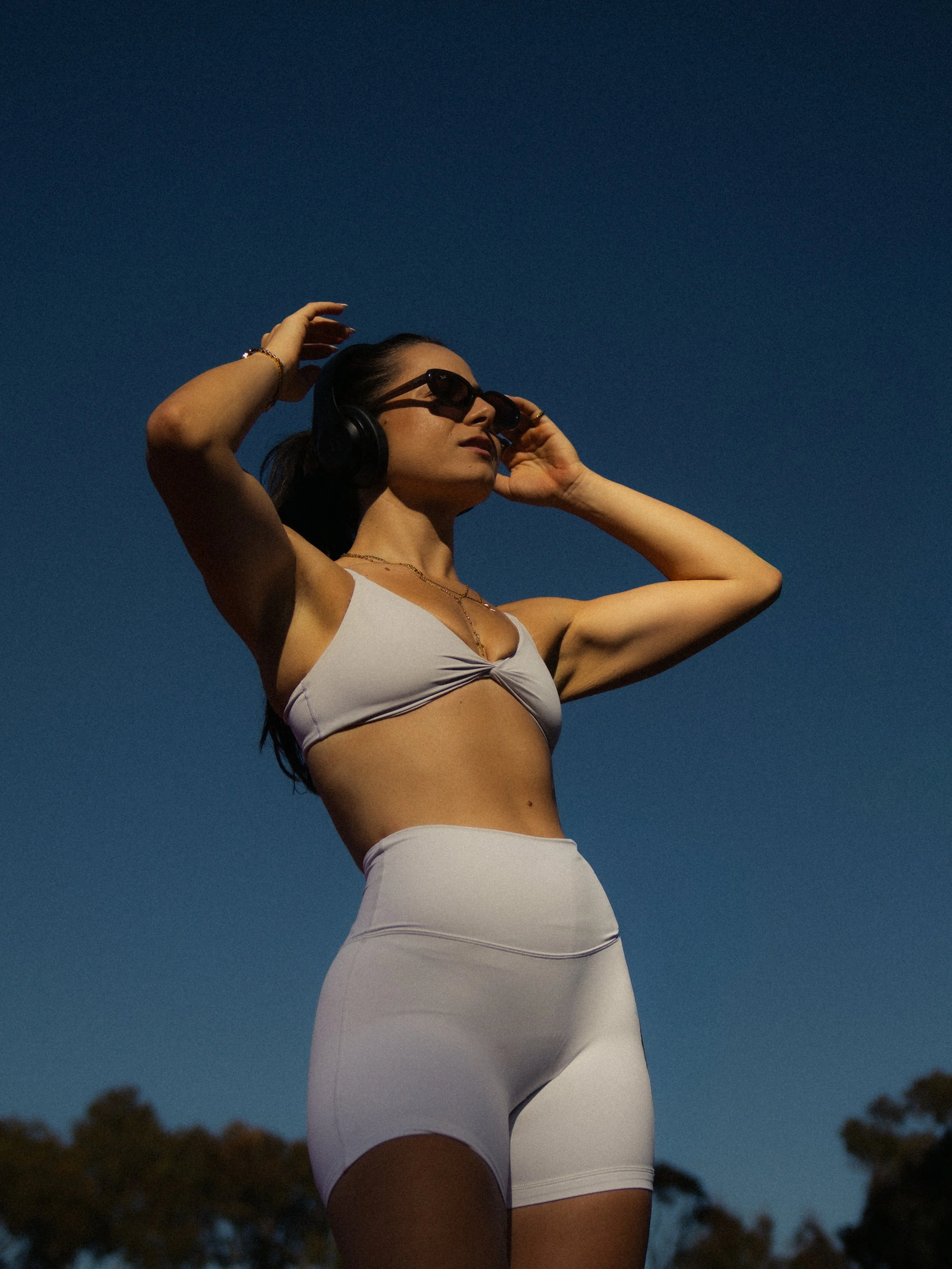 A woman wearing sunglasses, headphones, a white sports bra, and white leggings standing outdoors against a darkening sky, adjusting her headphones.