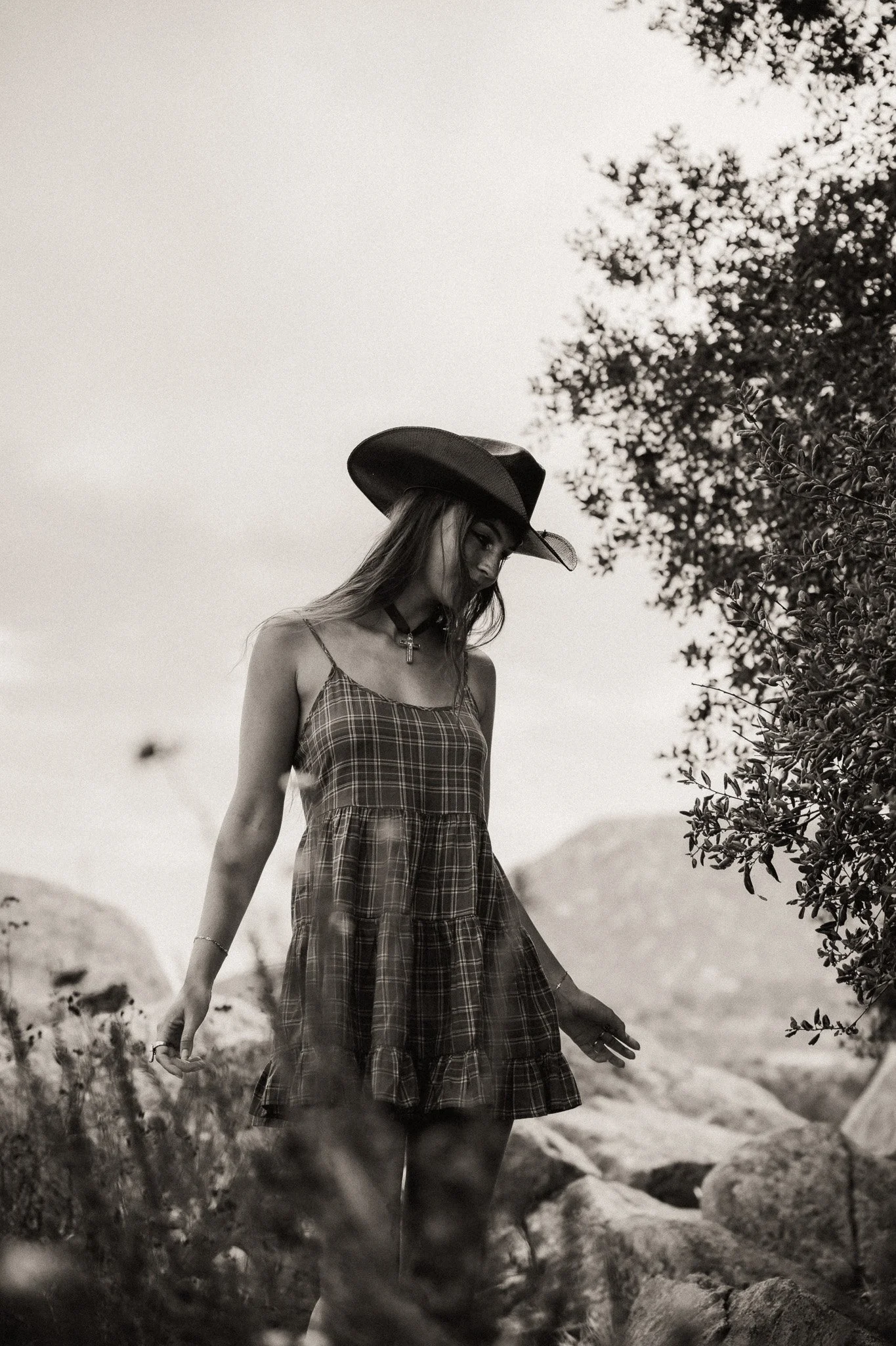 A woman in a plaid dress and cowboy hat walking outdoors on rocky terrain near bushes and trees, black and white photograph.