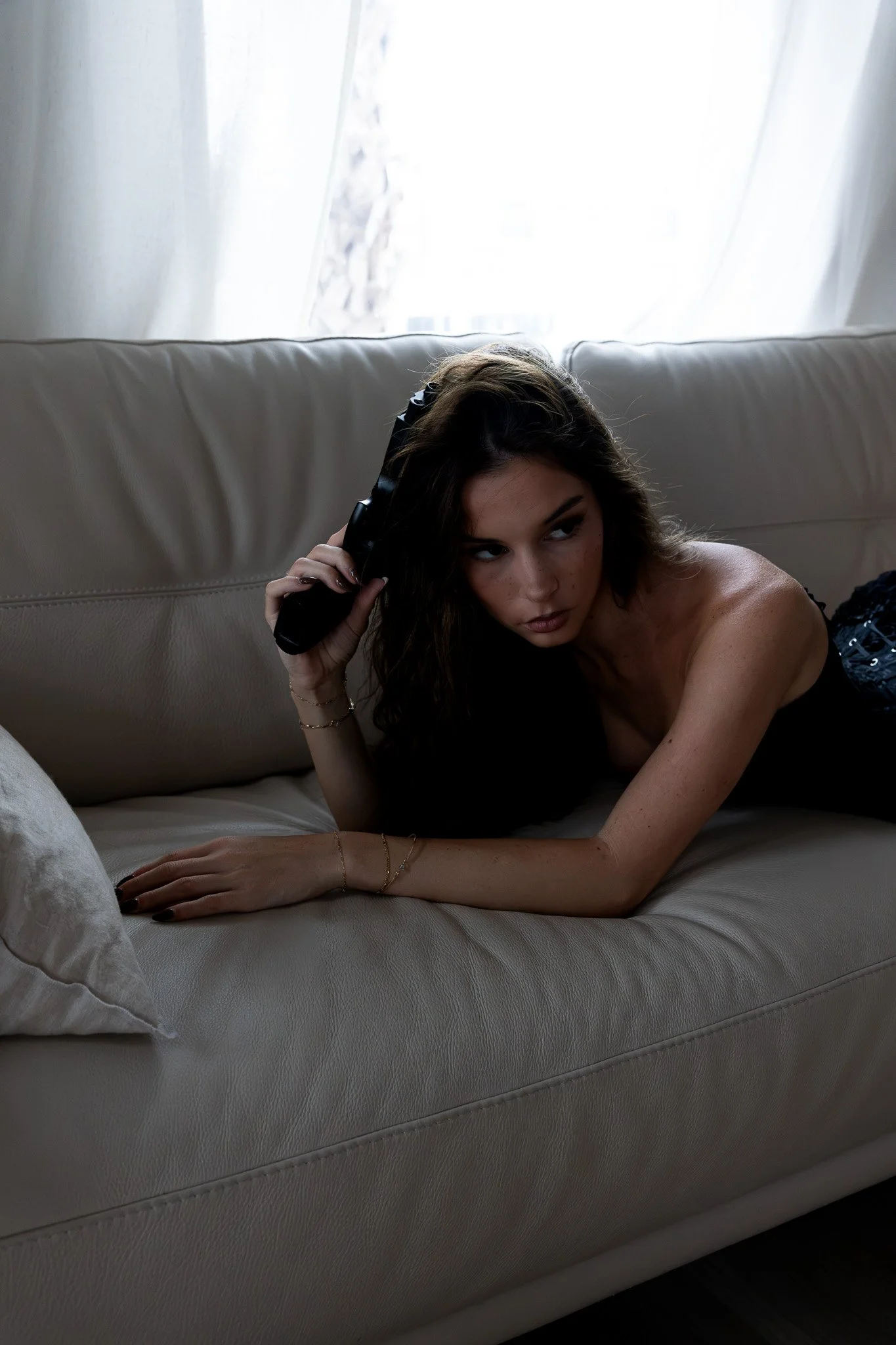 A woman with long dark hair lying on a beige couch, holding a black hairbrush to her head, with a sheer white curtain and window in the background.