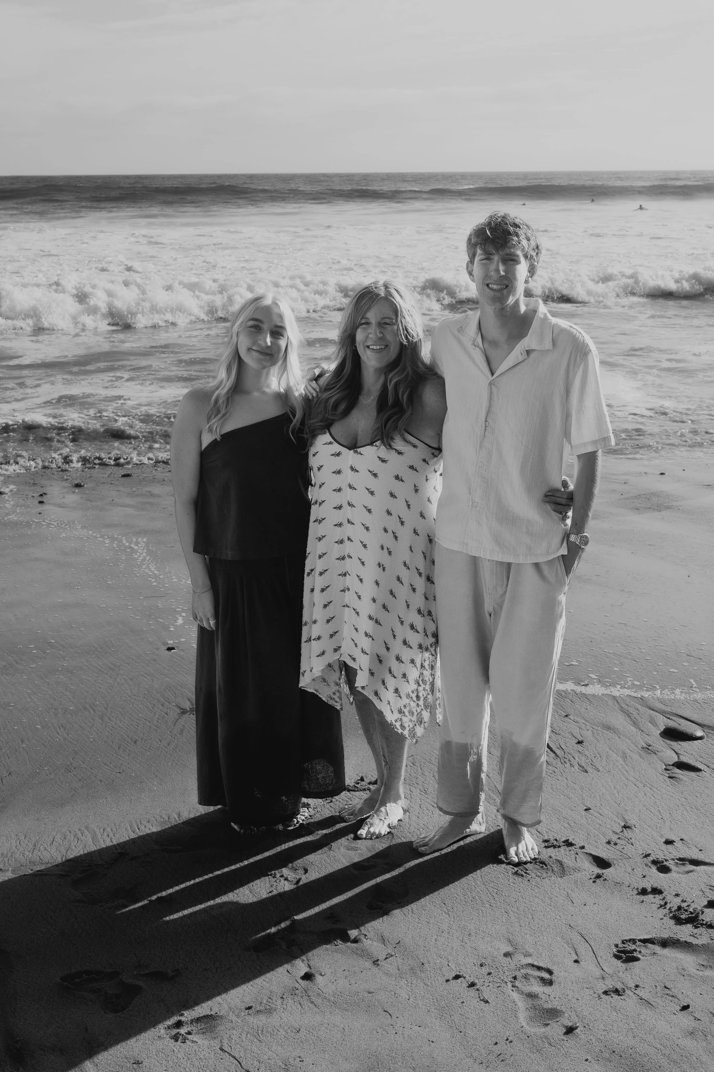 Black and white photo of three people hugging on a beach, smiling, with ocean waves in the background and surfboards in the distance.