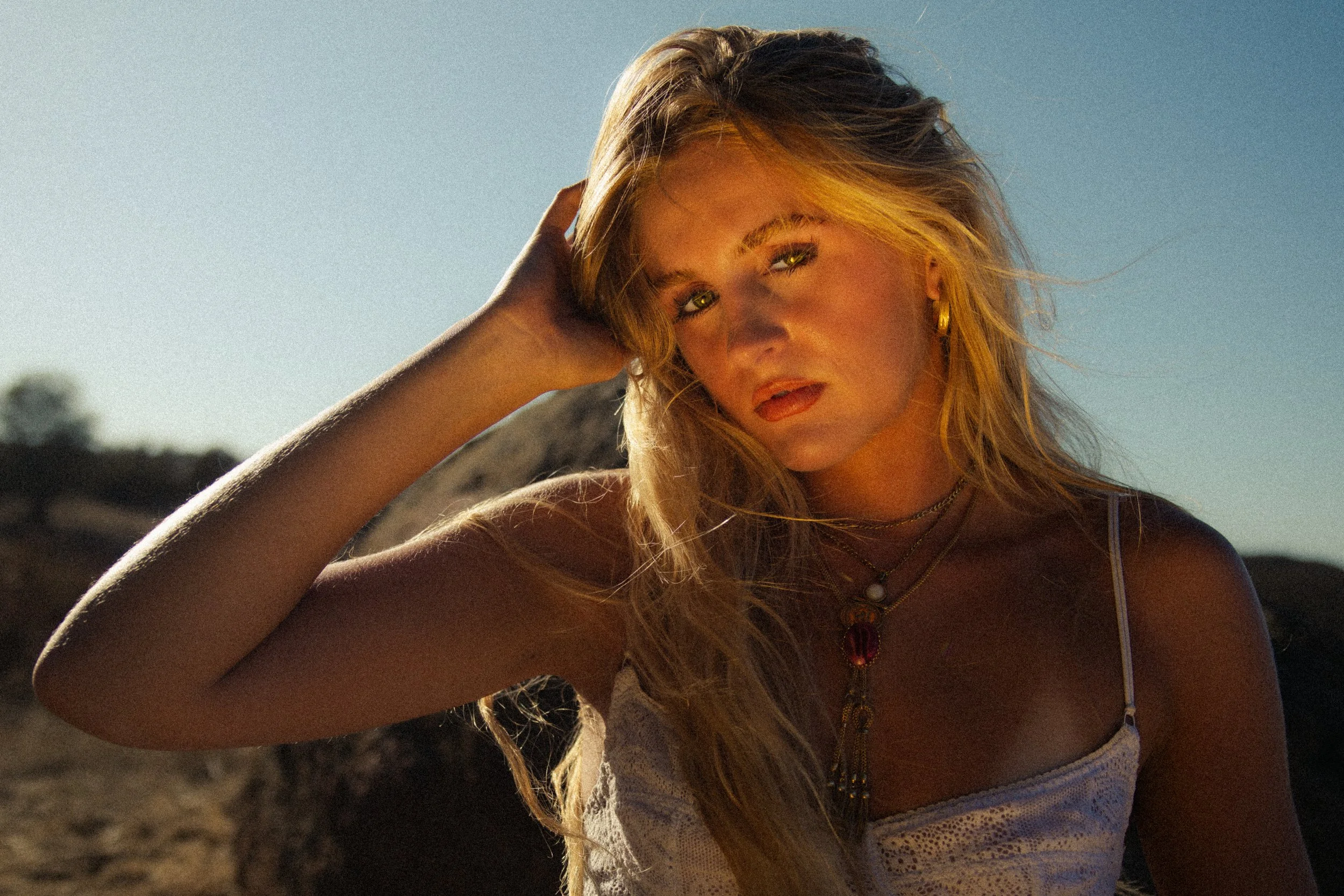 A woman with long, wavy blonde hair and golden eyes, wearing gold hoop earrings and layered necklaces, posing outdoors with a clear sky in the background.