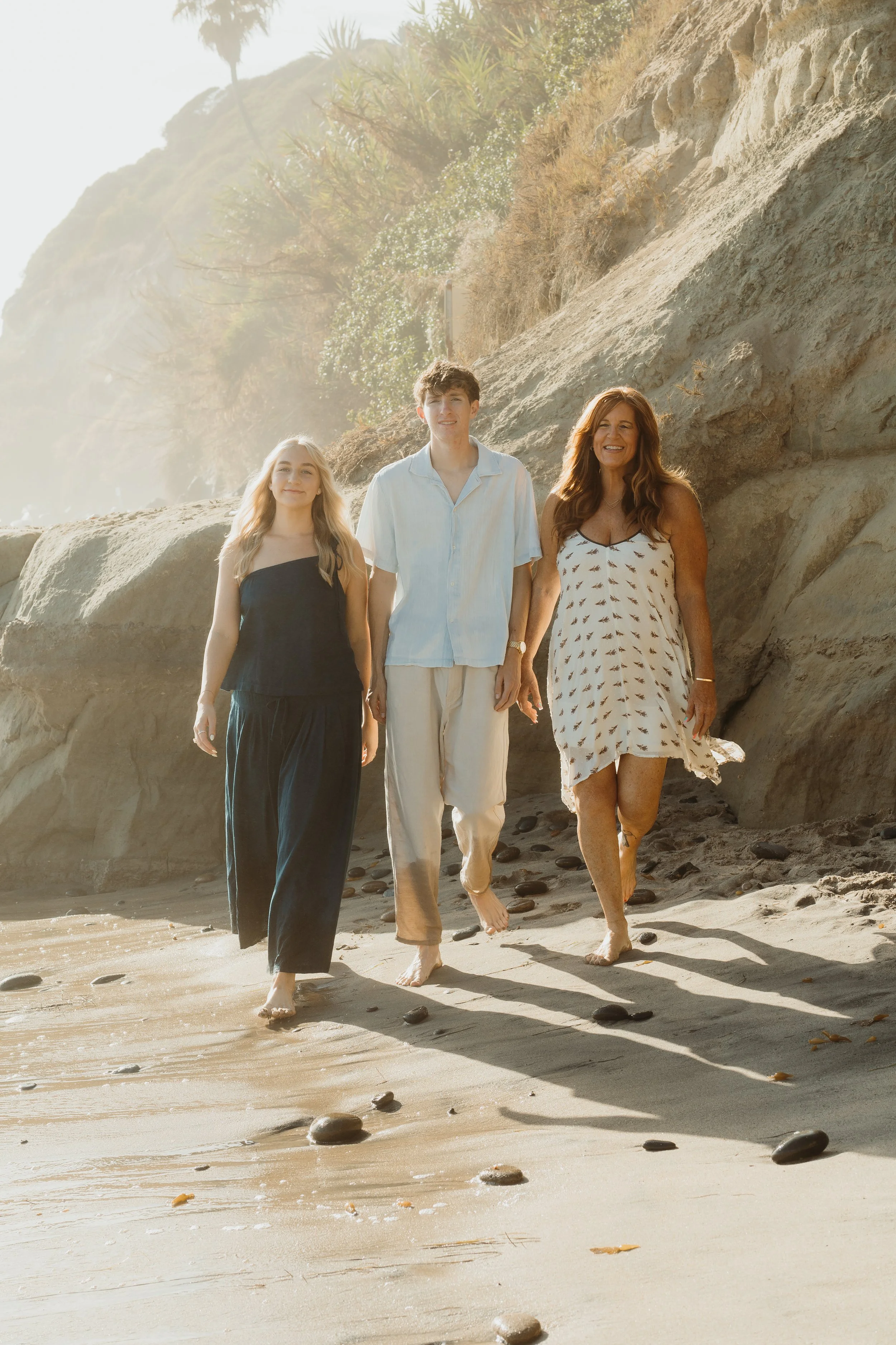 Three people walking barefoot on a sandy beach with rocks, with cliffs and trees in the background, during daylight.