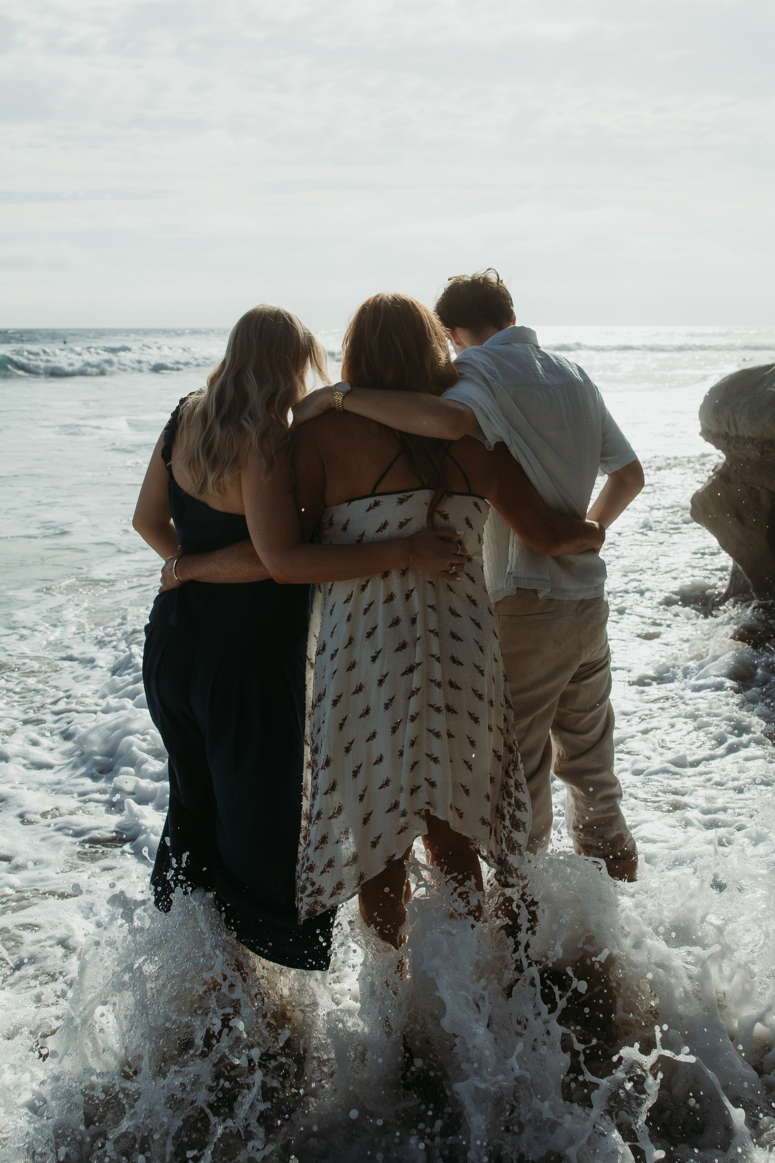 Three people walking in the ocean waves at the beach during daylight, seen from behind. One person has arm around another, with close hair and wedding ring visible. The third person has arm around the others, wearing a light-colored shirt. The sky is