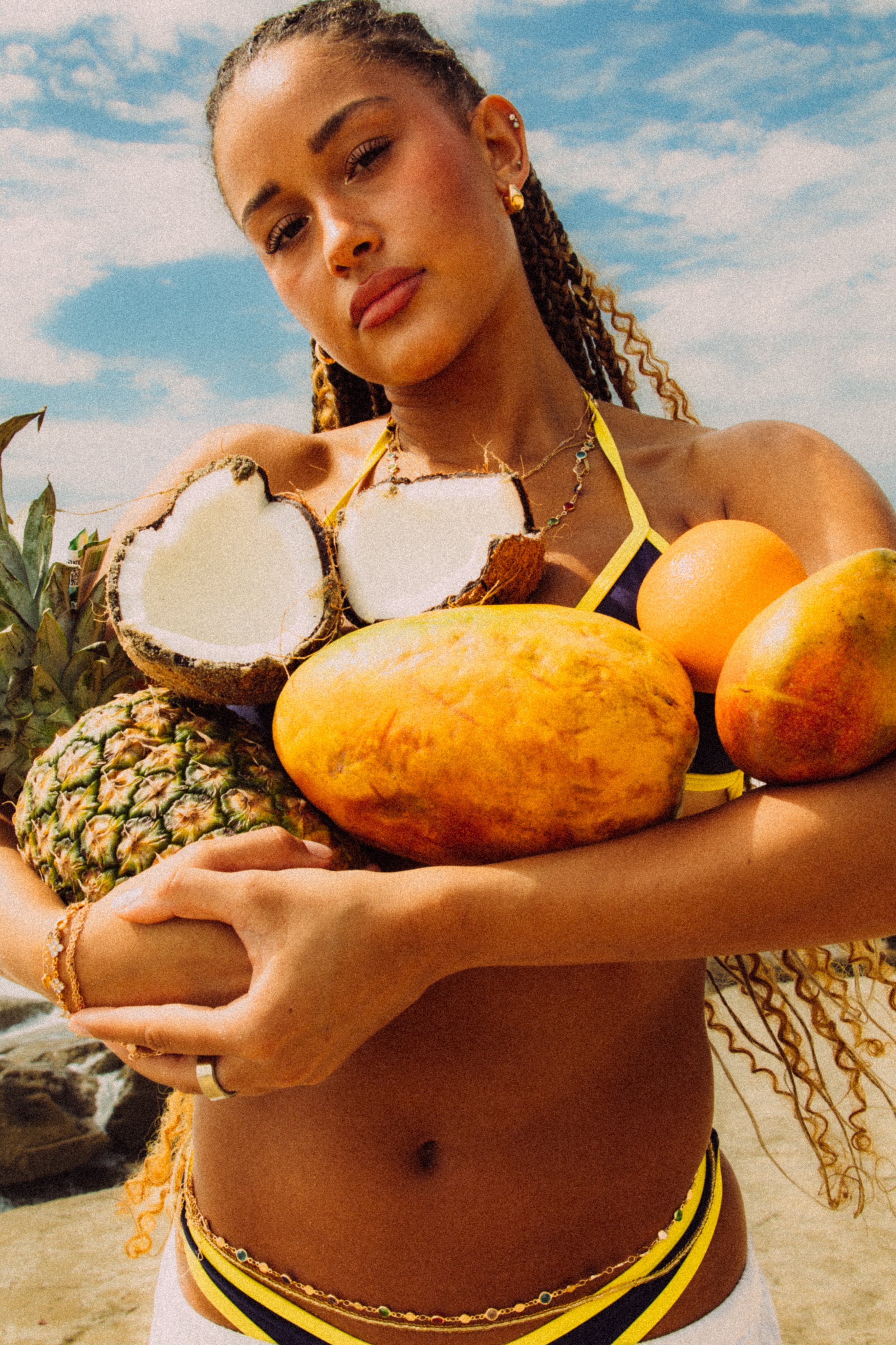 A woman with braided hair holds a variety of tropical fruits including a pineapple, a coconut, and mangoes, with a blue sky and clouds in the background.