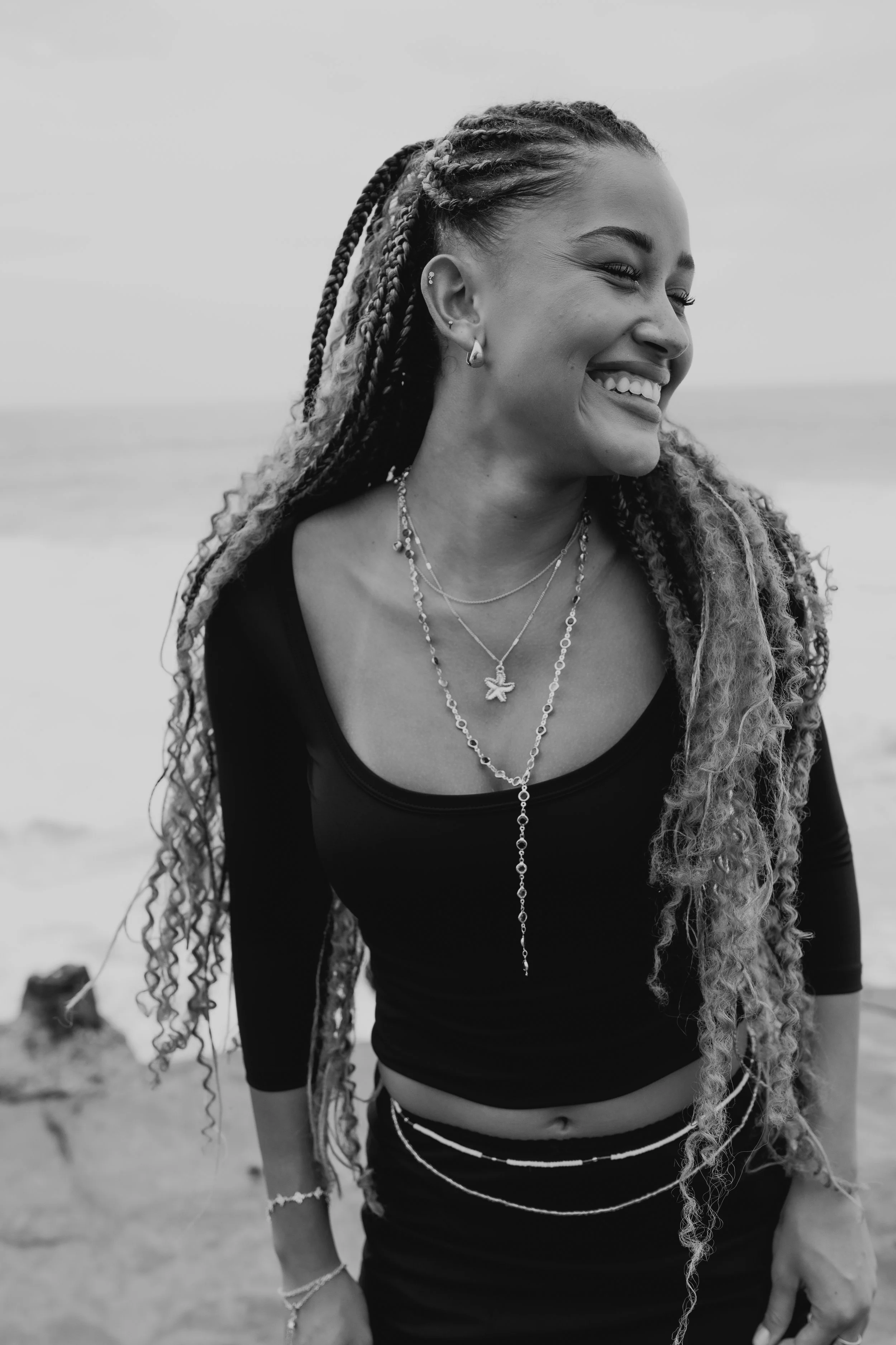 A smiling woman with braided hair and multiple necklaces on the beach, wearing a black top.