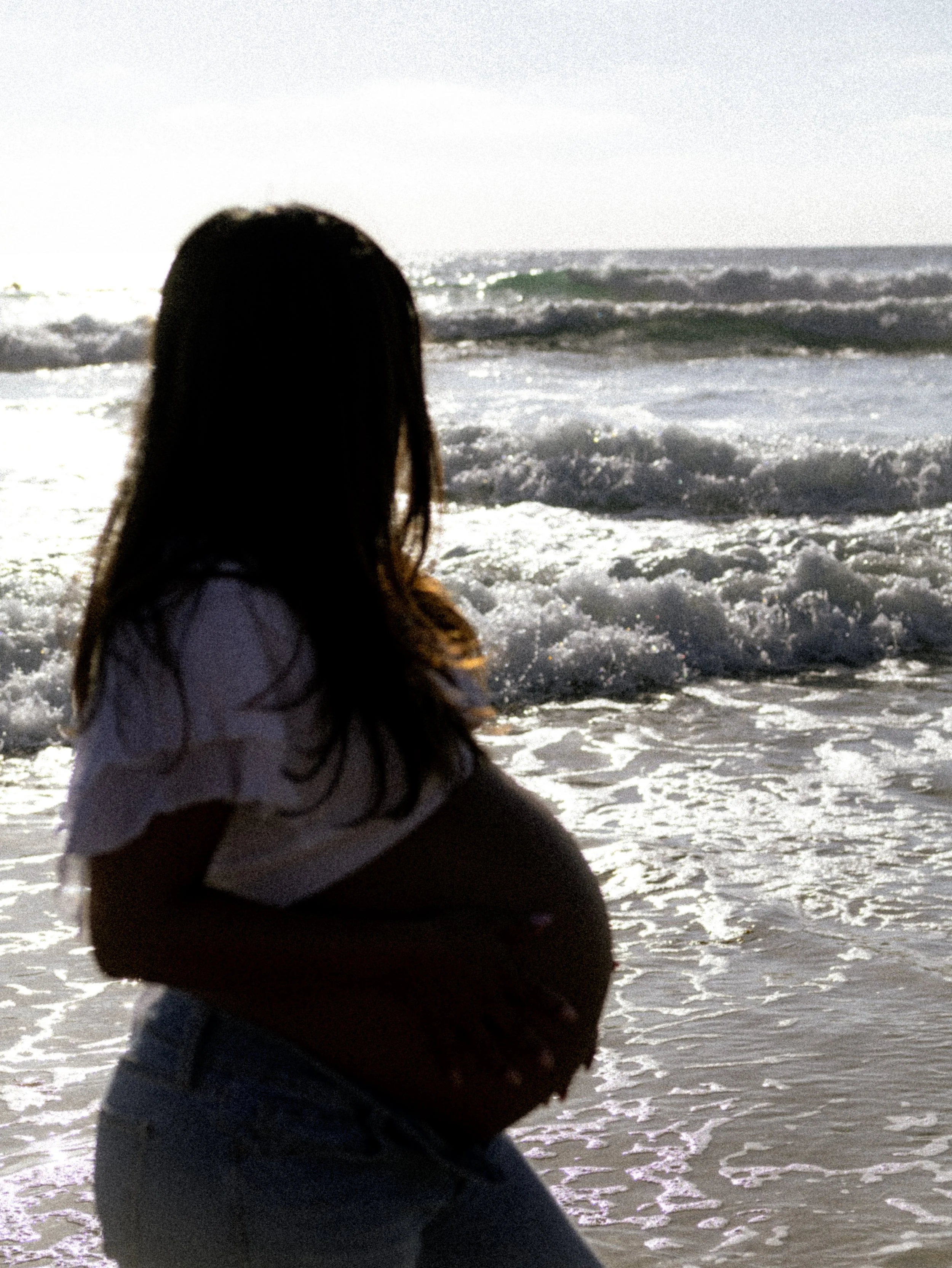 Person with long hair in a hoodie sitting on the beach, looking out at the ocean waves.