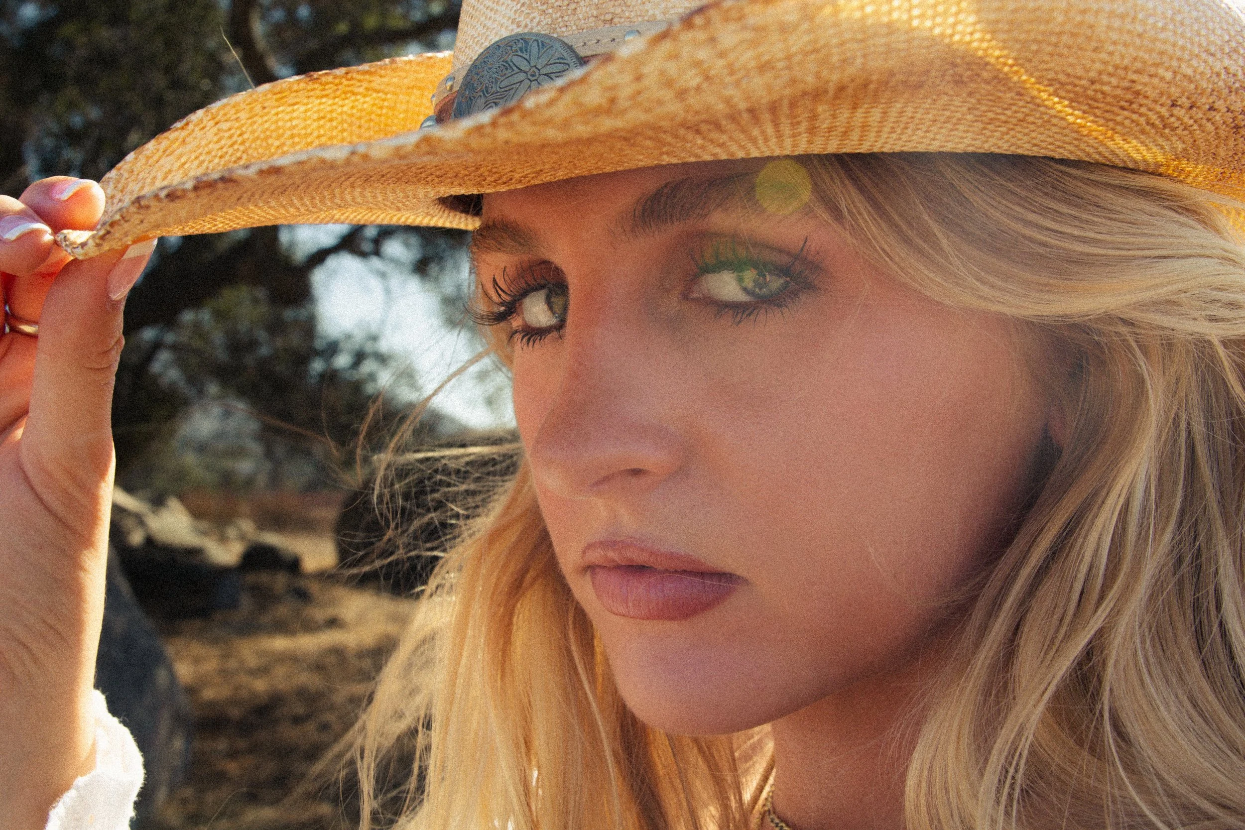 A woman with long blonde hair wearing makeup and a wide-brimmed straw hat, holding the brim of her hat with her left hand, outdoors with blurred trees in the background.