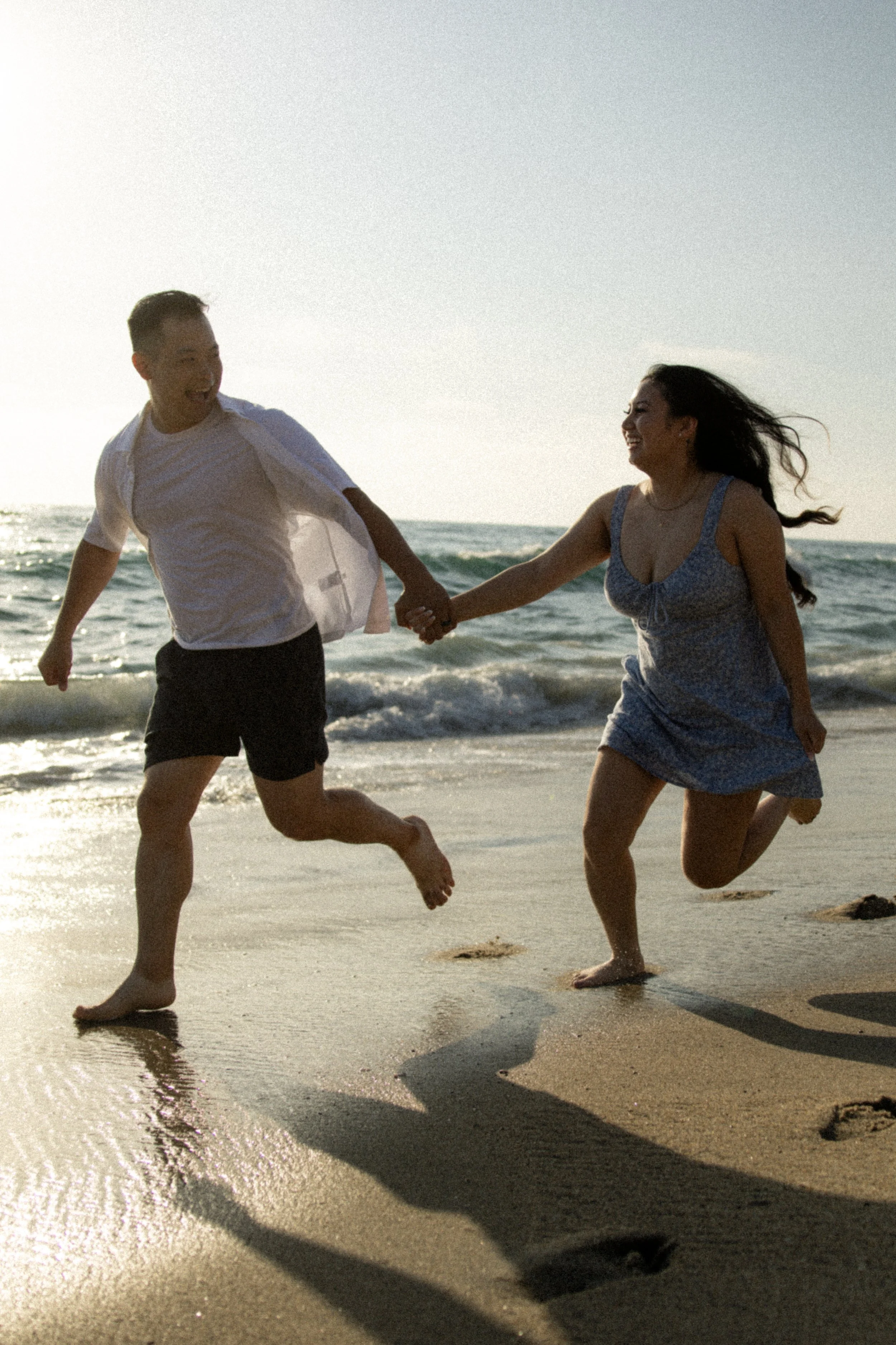 Two people, a man and a woman, running and holding hands on a beach with the ocean waves in the background during sunset.