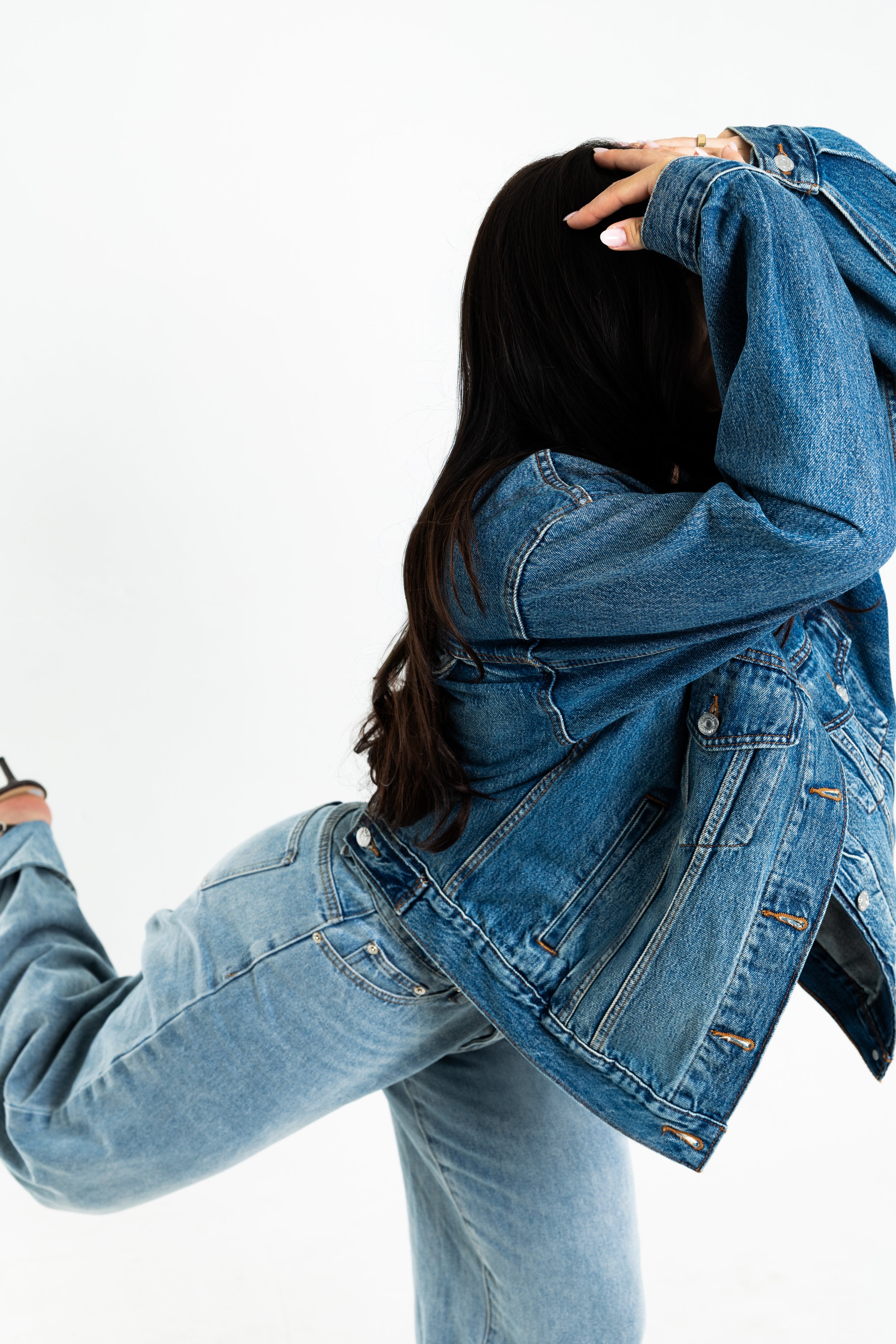 Person wearing a denim jacket and light blue jeans, holding their head with both hands, in a studio with a white background.