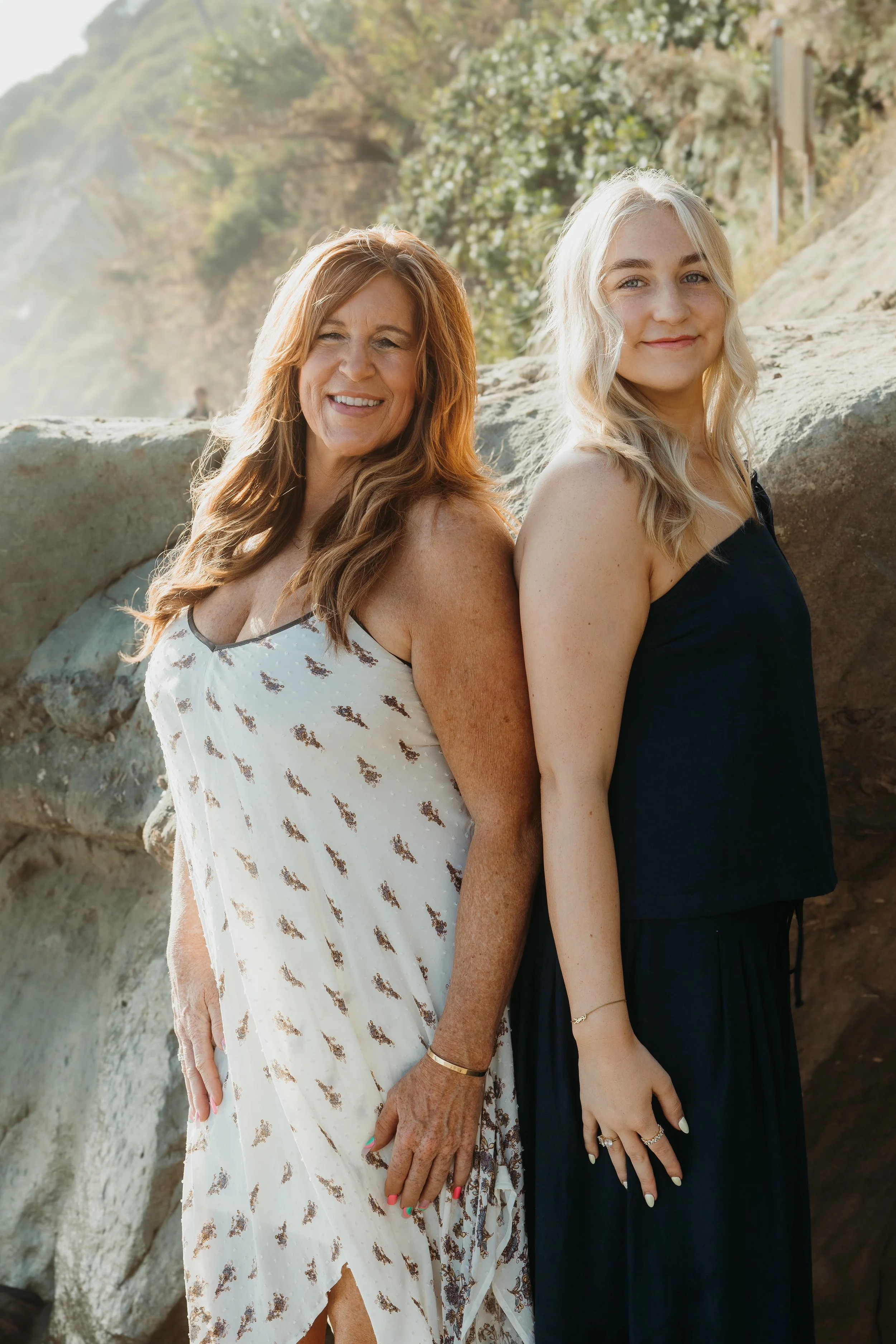 Two women standing in front of rocks with greenery in the background, smiling at the camera. One has red hair and is wearing a white dress with a pattern, the other has blonde hair and is wearing a black dress.