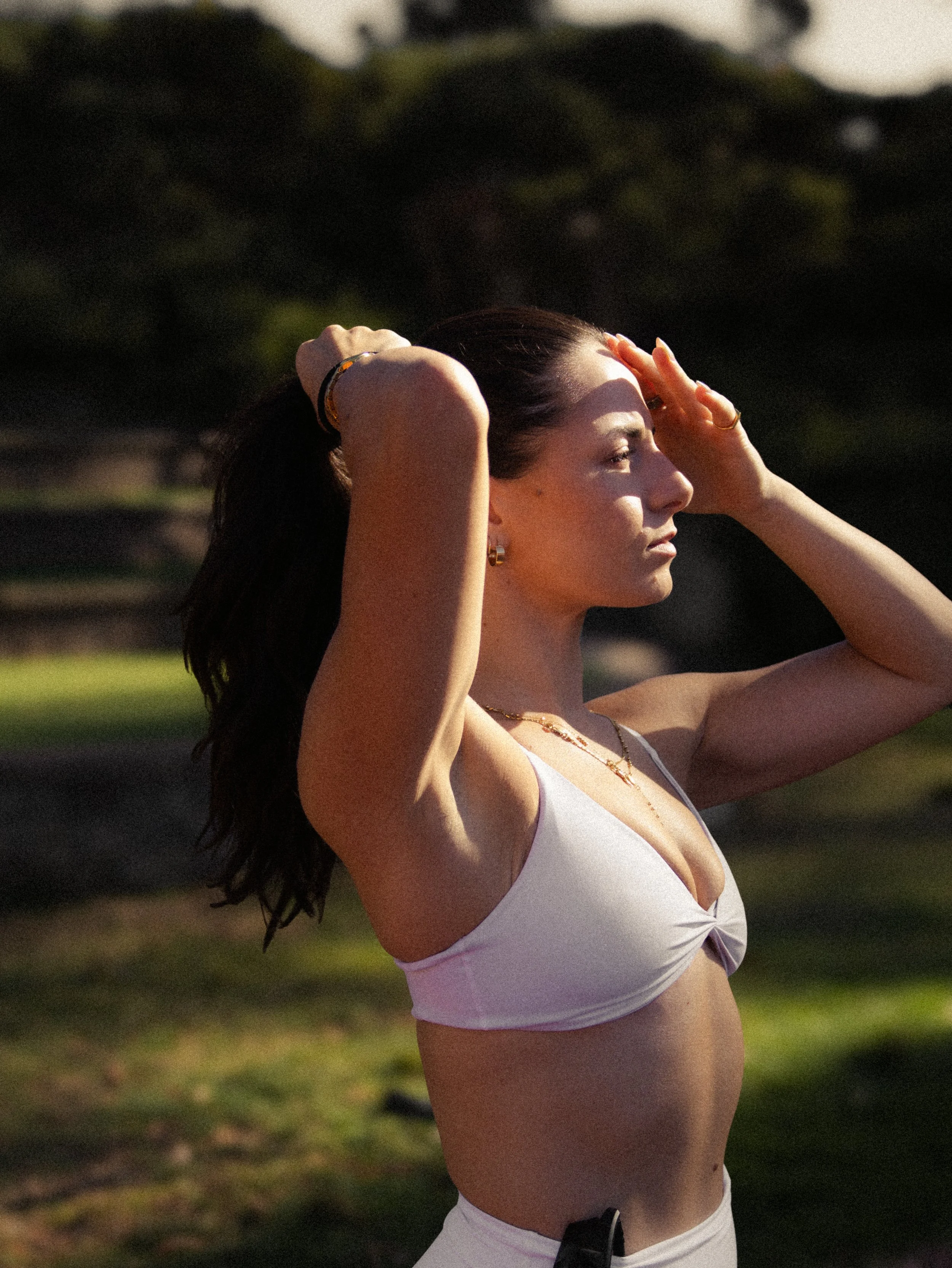 A woman with long dark hair and tan skin, wearing a white sports bra and jewelry, standing outdoors with her hands on her forehead, with her eyes closed and a serene expression, in a park or garden setting during sunset.