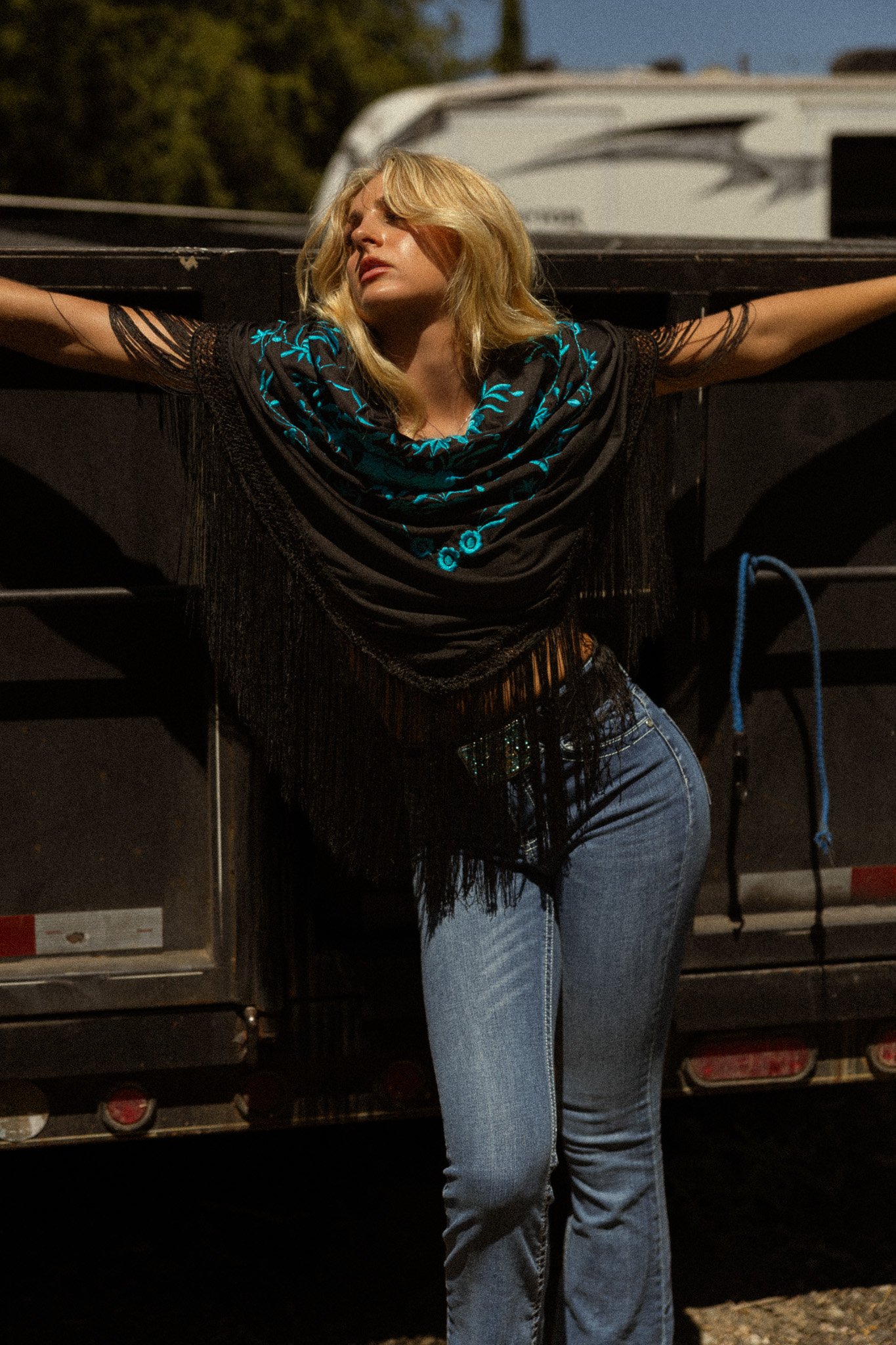 A woman with blonde hair wearing a black top with turquoise embroidery and blue jeans standing with arms outstretched against a black truck at sunset.