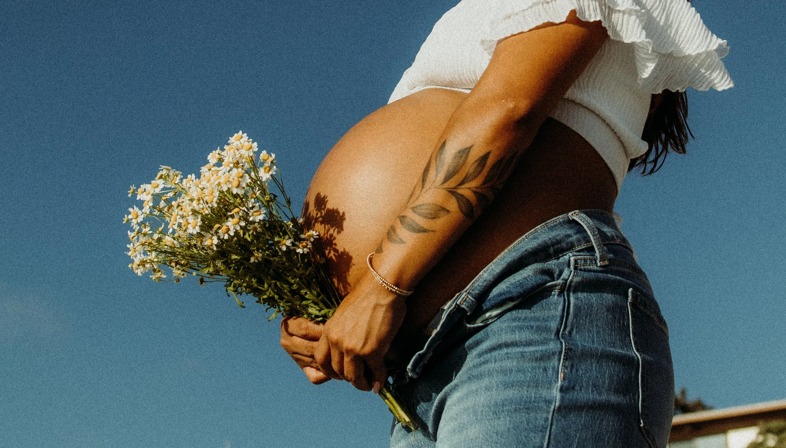 A person wearing a white off-shoulder top and jeans is holding a bouquet of daisies against a blue sky background.