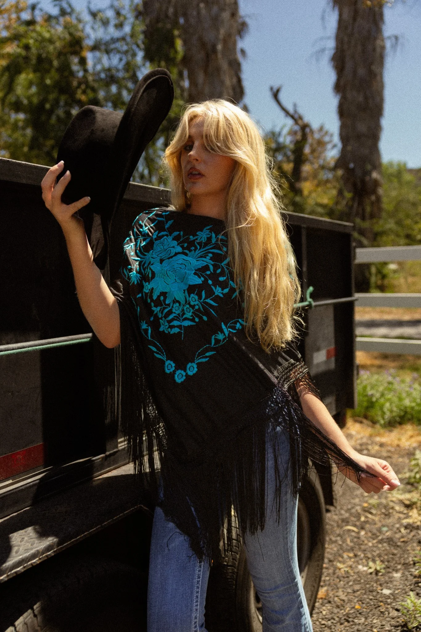 A woman with long blonde hair wearing a black top with blue floral embroidery, holding a black hat, standing next to a black vehicle with an open door, outdoors with trees and clear sky in the background.