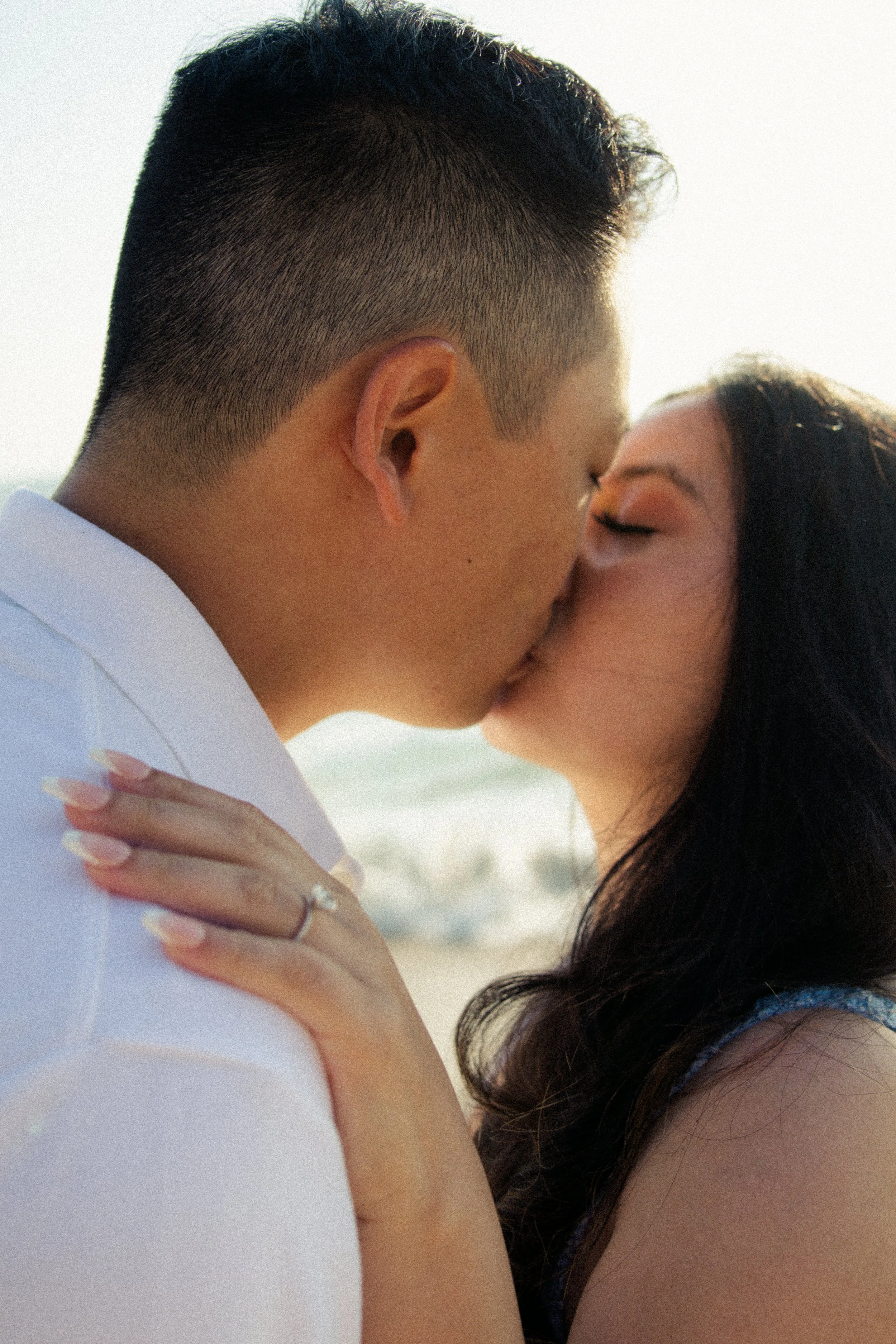A couple about to kiss outdoors, with the man's face and part of the woman's face visible. The woman has her eyes closed and is resting her hand on the man's shoulder, showing a wedding ring.