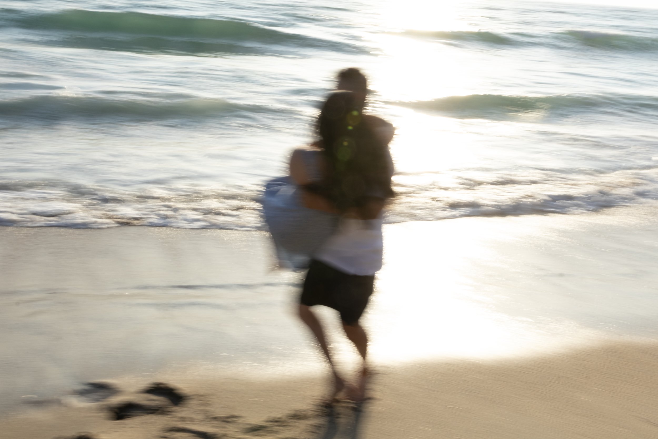 A person running or walking along the beach near the water with waves and sunlight reflecting on the surface.