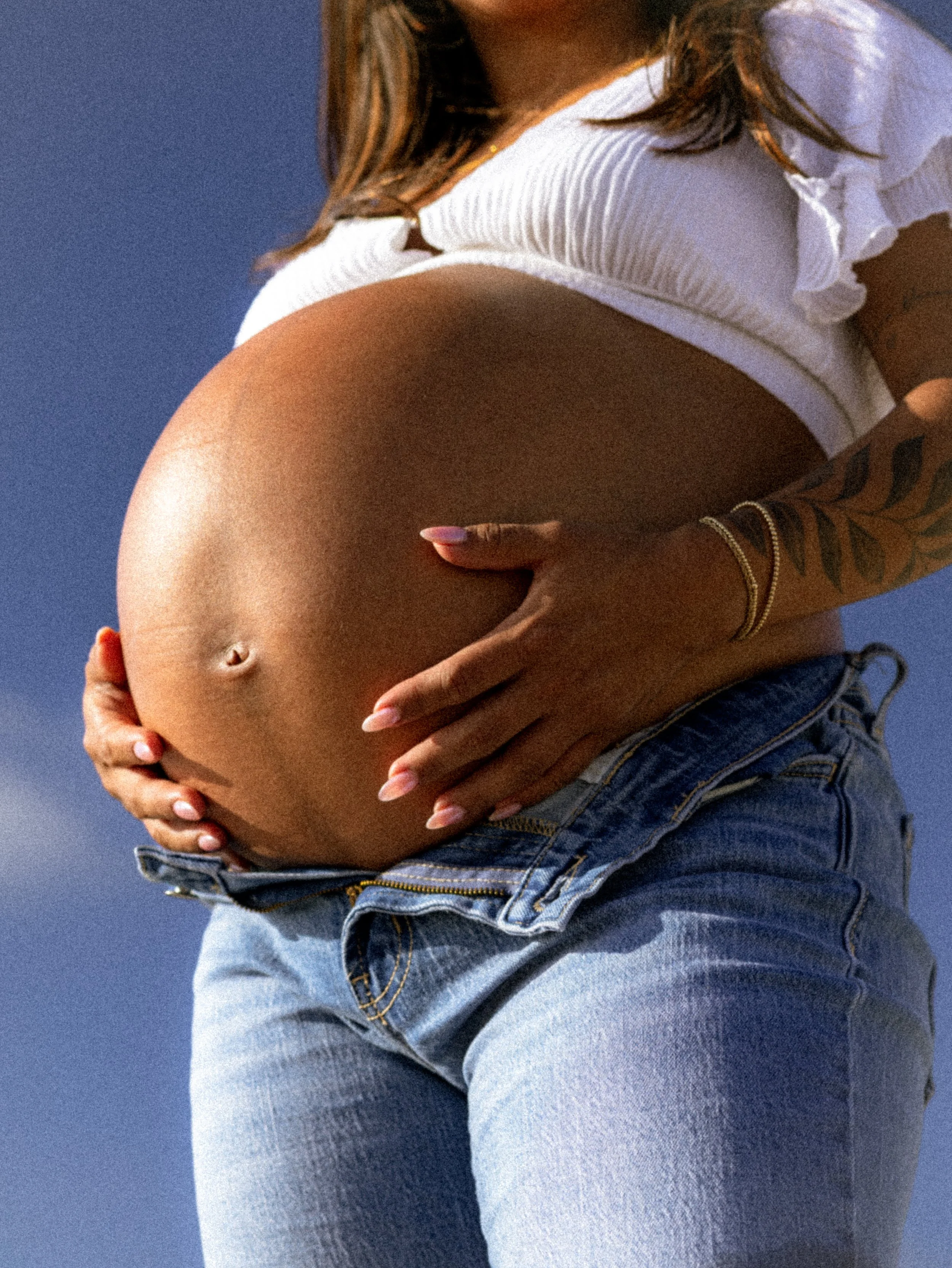Close-up of a woman with a tanned belly, wearing a white cropped top and blue jeans, standing in front of a blue background.