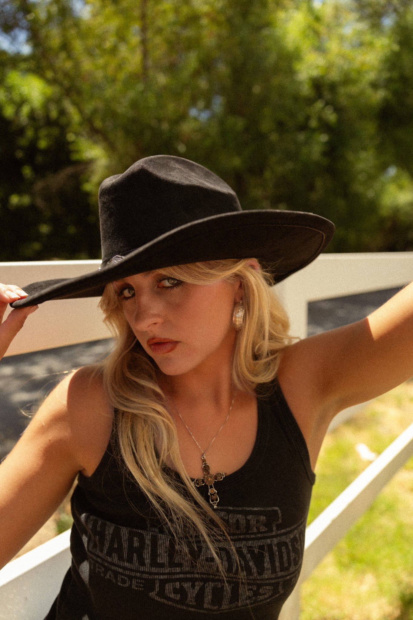 A woman with long blonde hair wearing a large black hat, black tank top, and jewelry, holding the brim of her hat while taking a selfie outdoors with trees in the background.