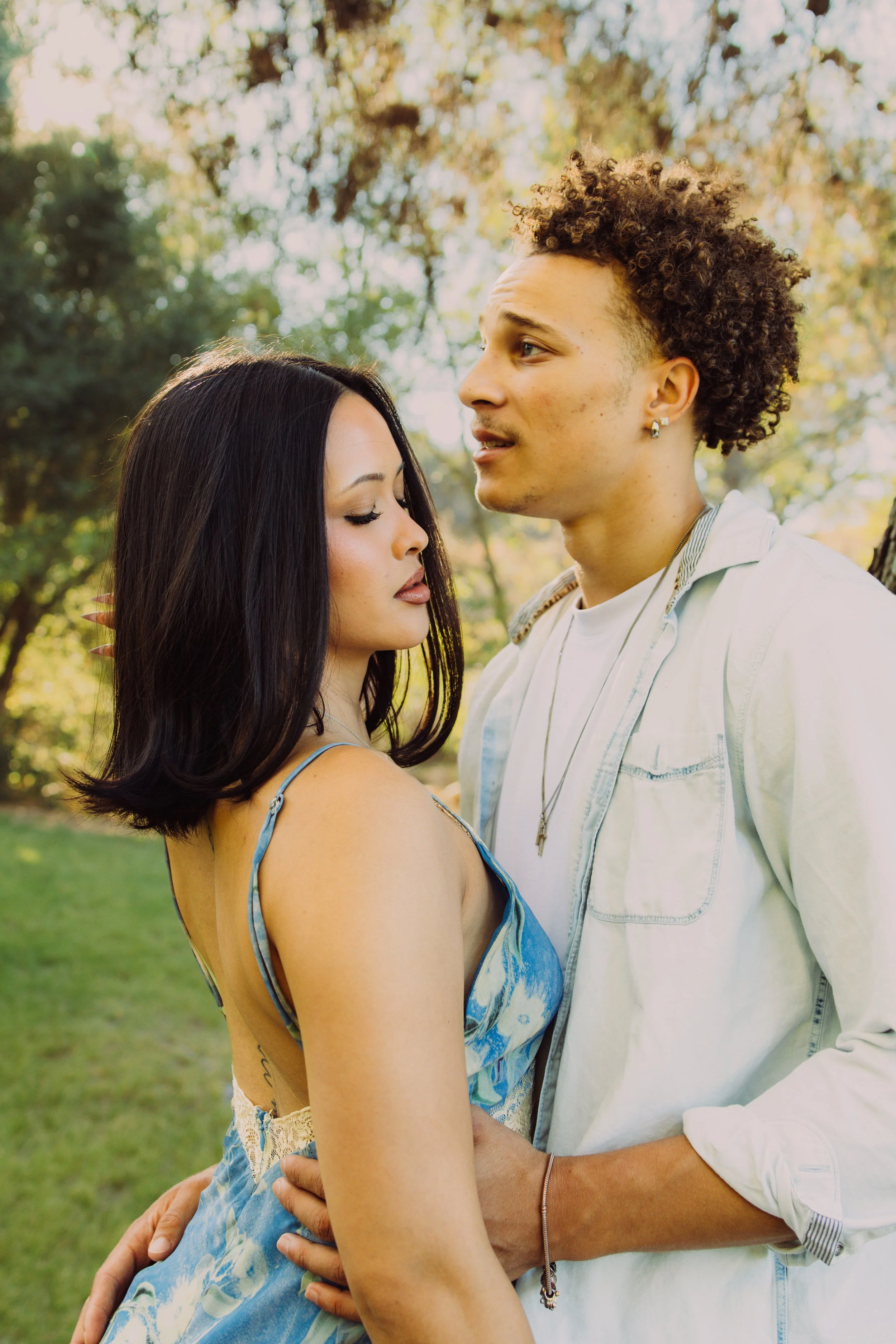 A young couple embracing outdoors in a park with sunlight filtering through trees, the woman with long black hair and the man with curly hair and earrings.