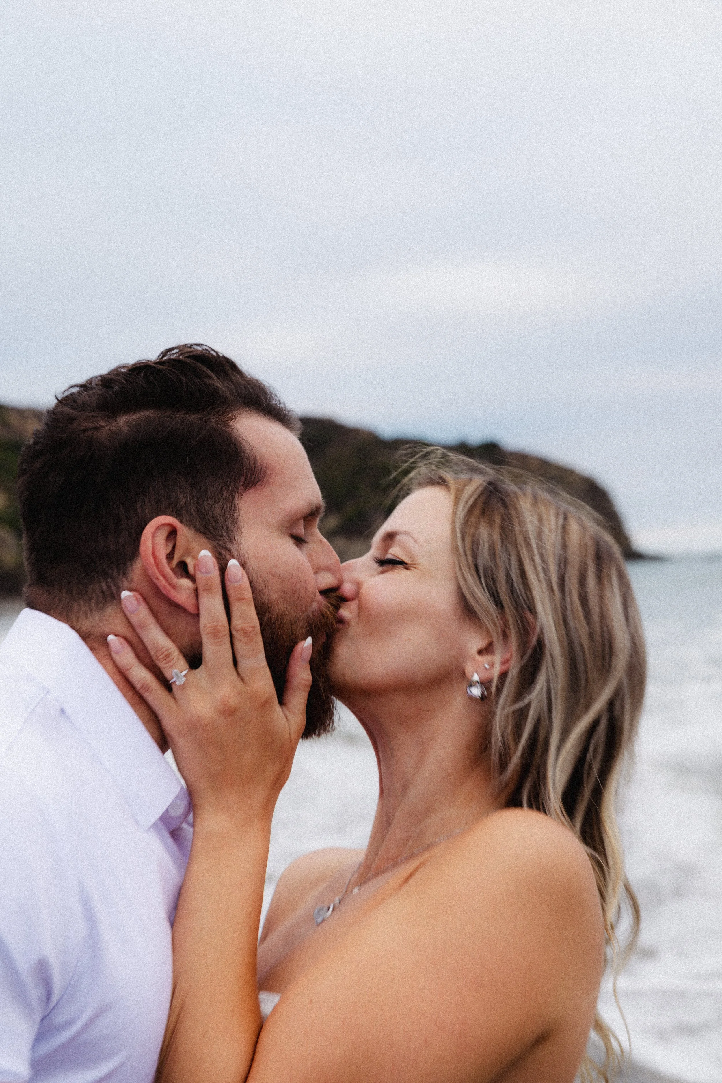 A couple passionately kissing outdoors with a beach and rocky hill in the background during daytime.