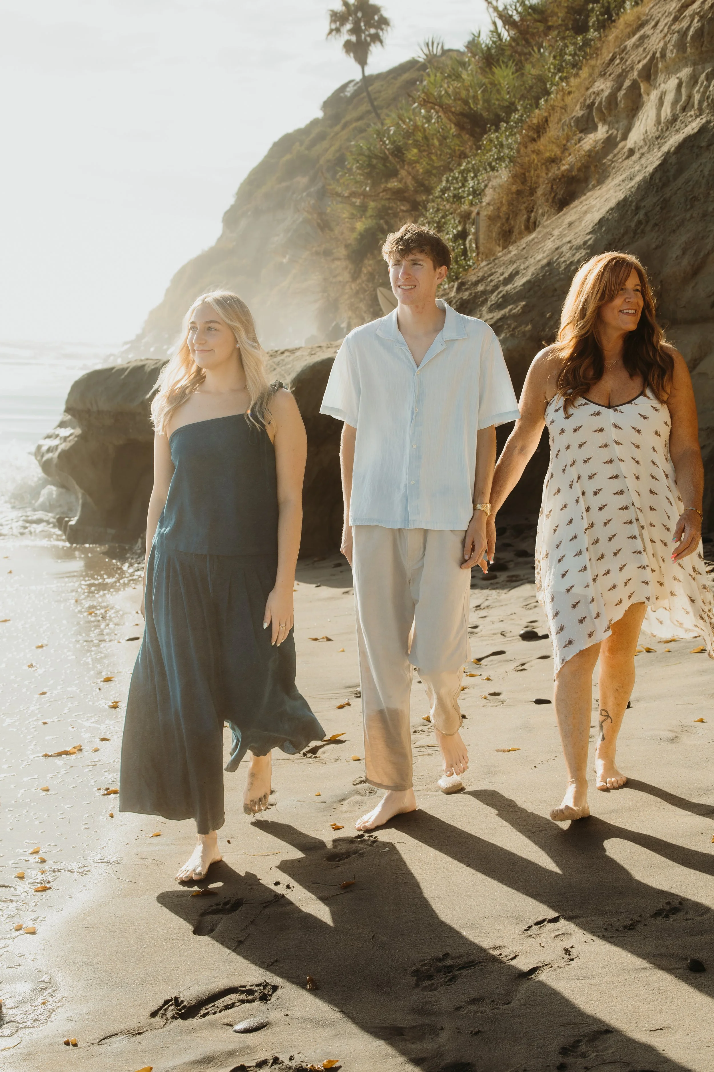 A group of four people, two women and two men, walking barefoot on a sandy beach with rocks and ocean in the background during the day. The scene is sunny with clear weather.