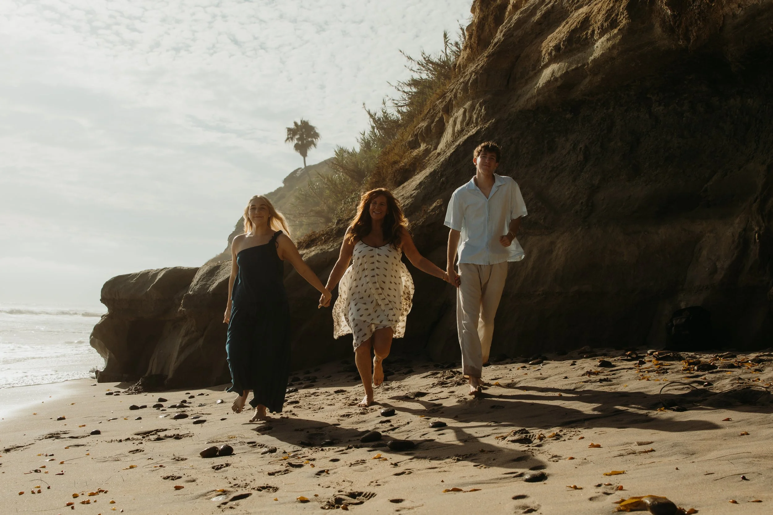 Three people walking along the beach near rocky cliffs with palm trees, in bright sunlight.