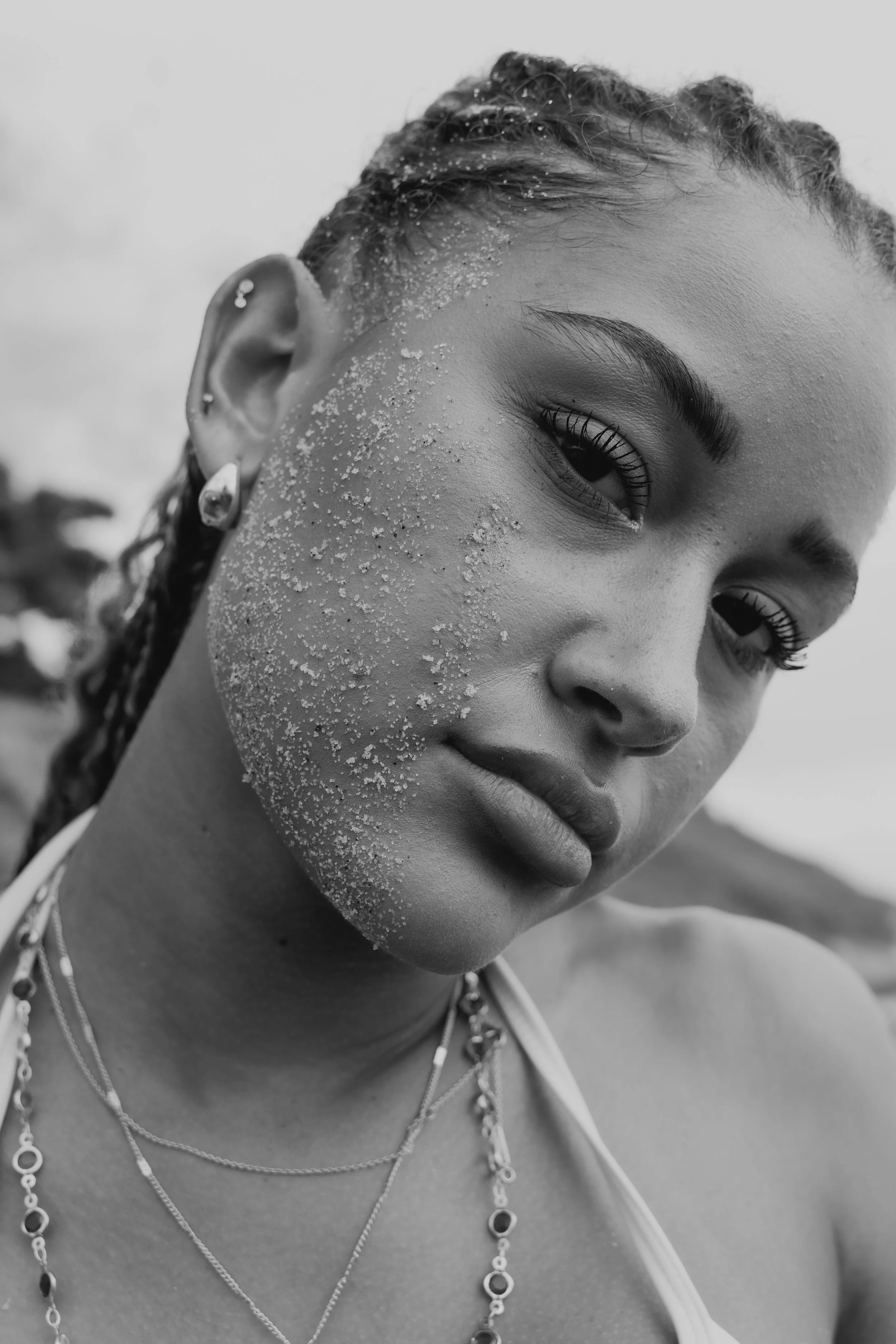Close-up of a young woman with sandy salt on her face, wearing multiple layered necklaces and earrings, with a blurred background.
