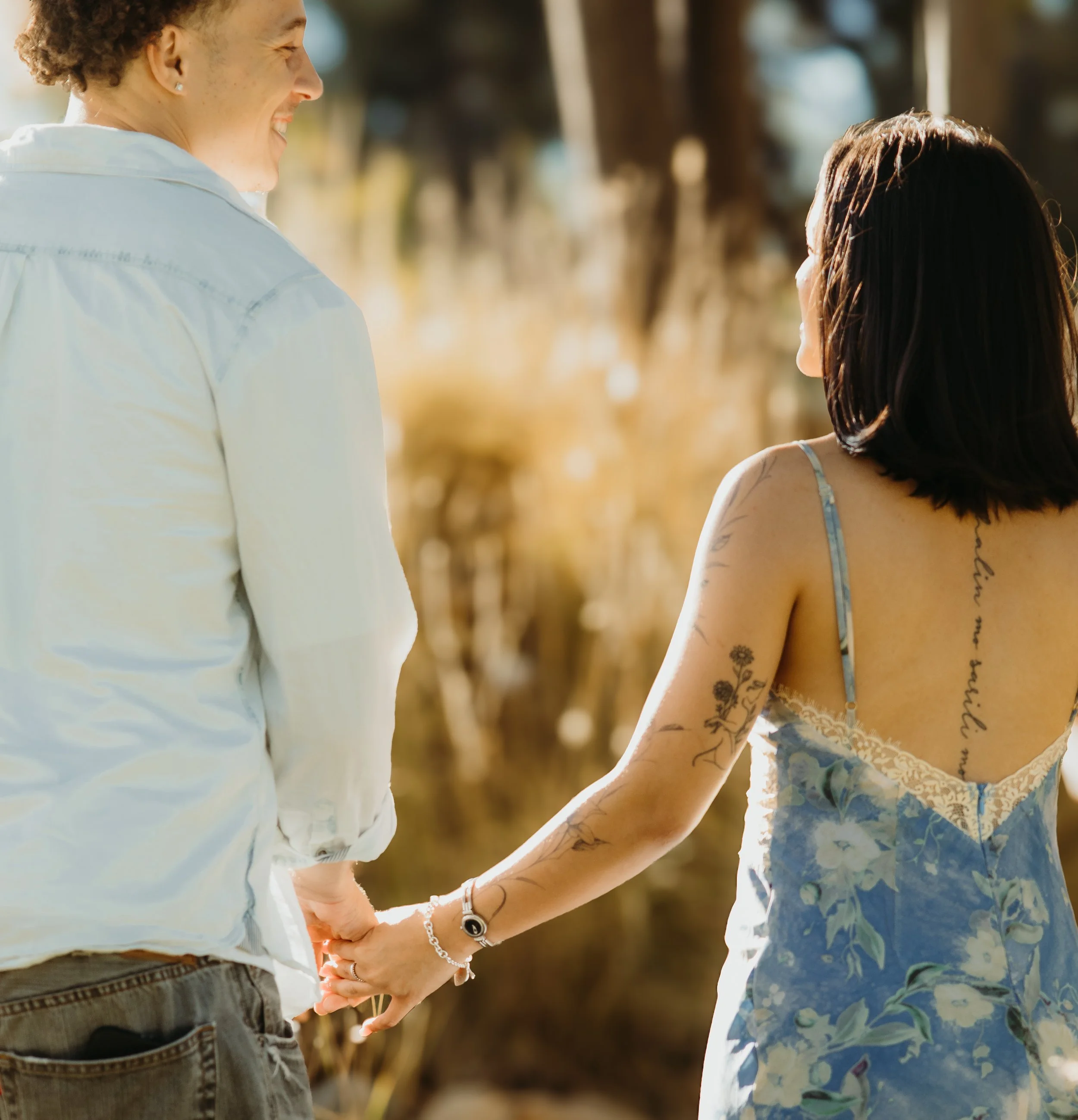 A man and a woman holding hands outdoors in a park during daytime, with trees in the background and sunlight illuminating their faces.