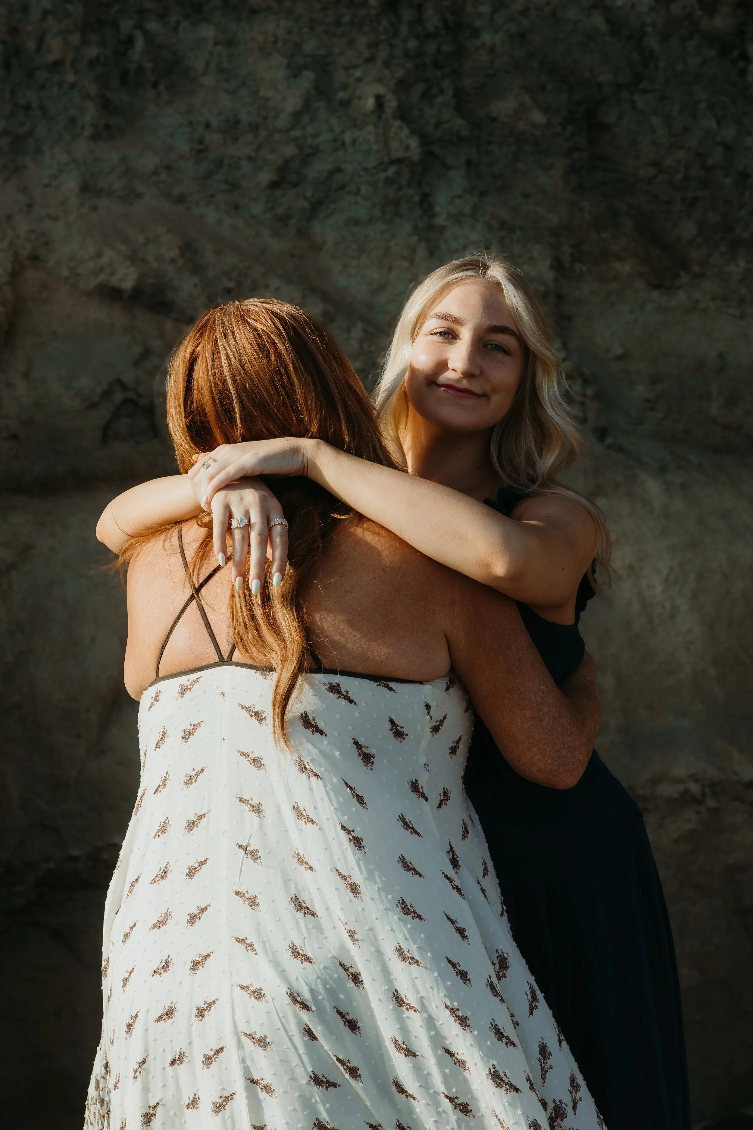 Two women embracing each other, one is wearing a white dress with a floral pattern and the other is in a black dress. They are outdoors with a rocky background, and sunlight is illuminating their faces.