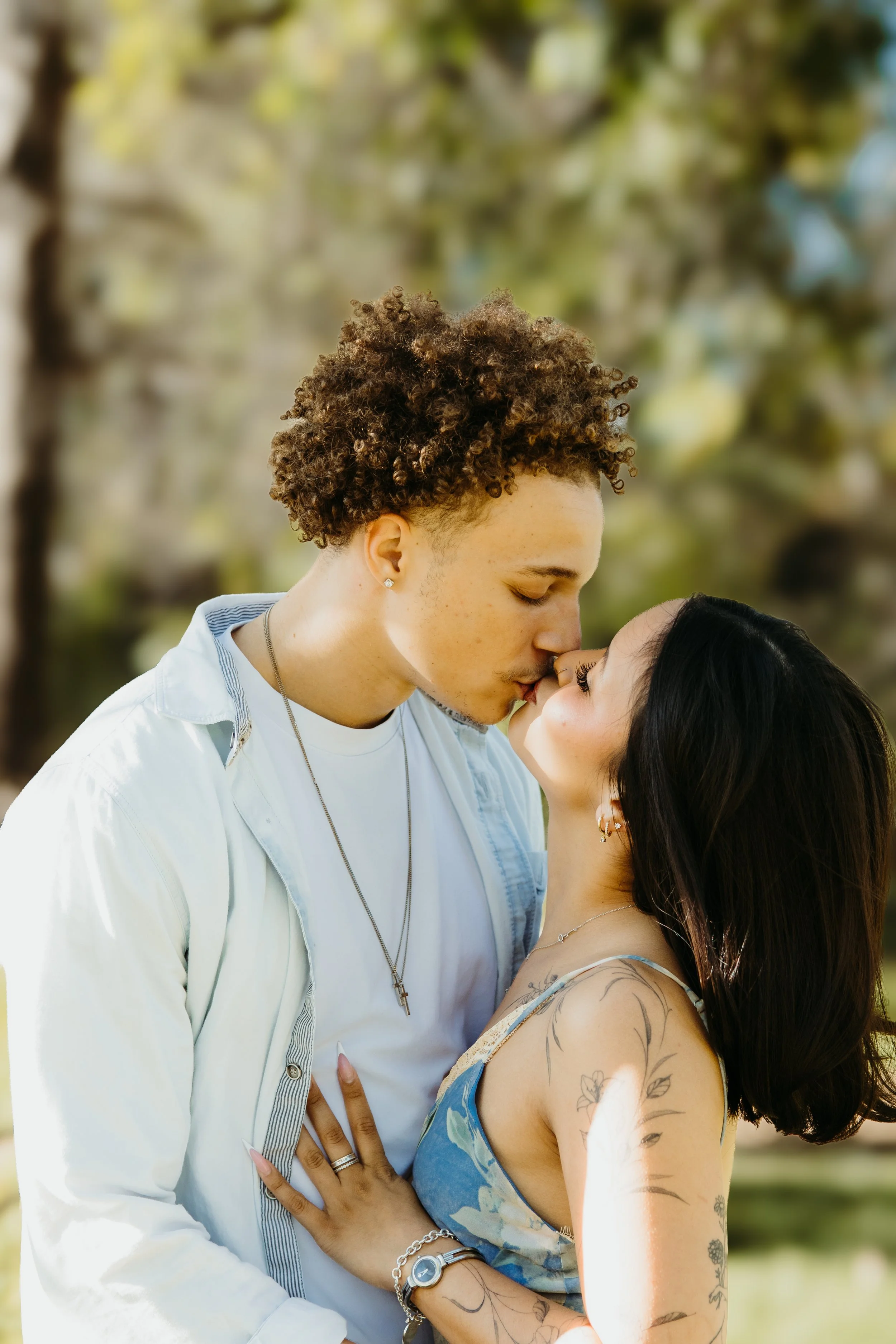 A couple sharing a tender moment outdoors, with the man gently kissing the woman's nose as she looks up at him, surrounded by a blurred green background.