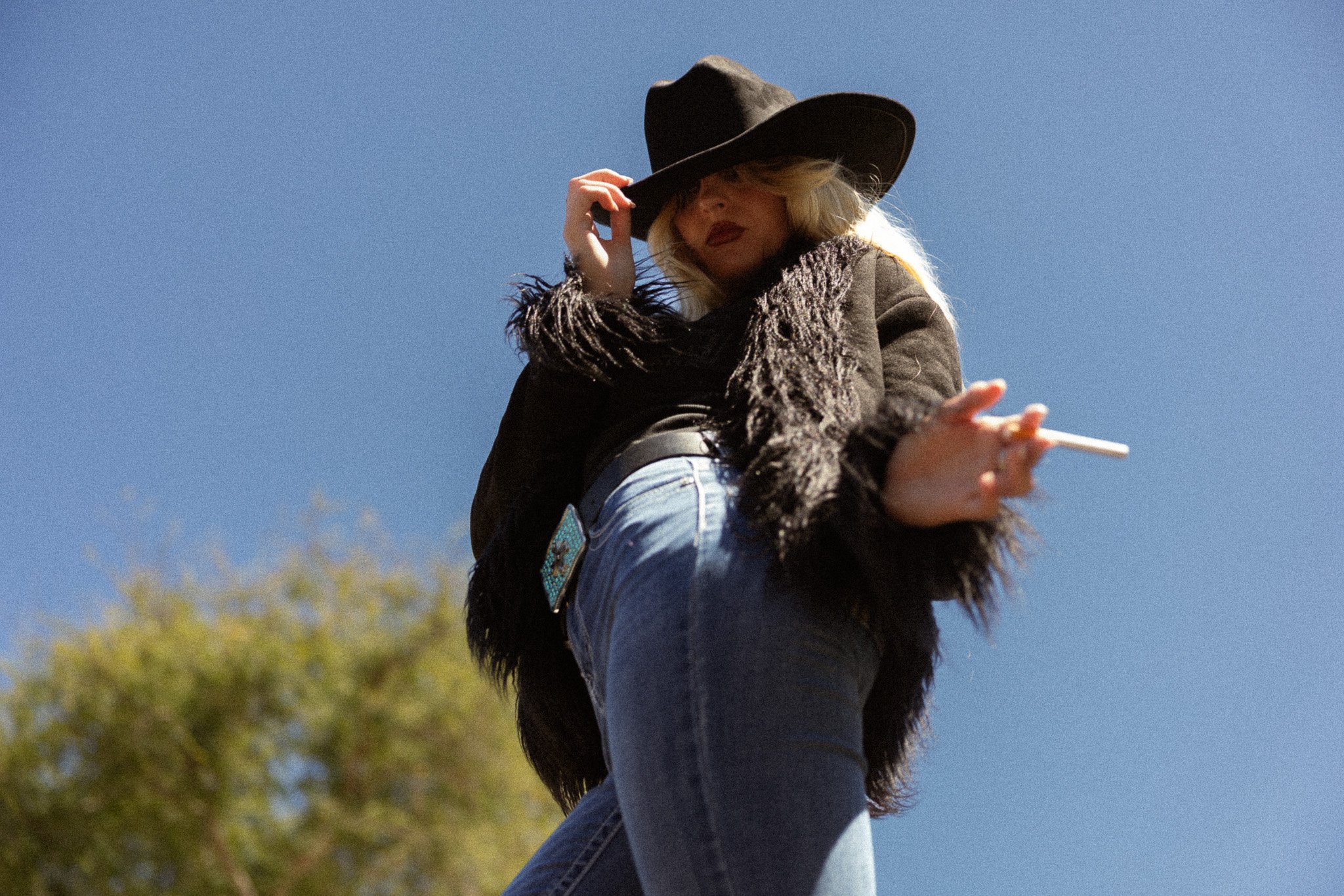 A woman with long blonde hair, wearing a wide-brimmed hat and a furry brown jacket, is sitting on the back of a horse, holding a cigarette in her hand, against a clear blue sky and green trees in the background.
