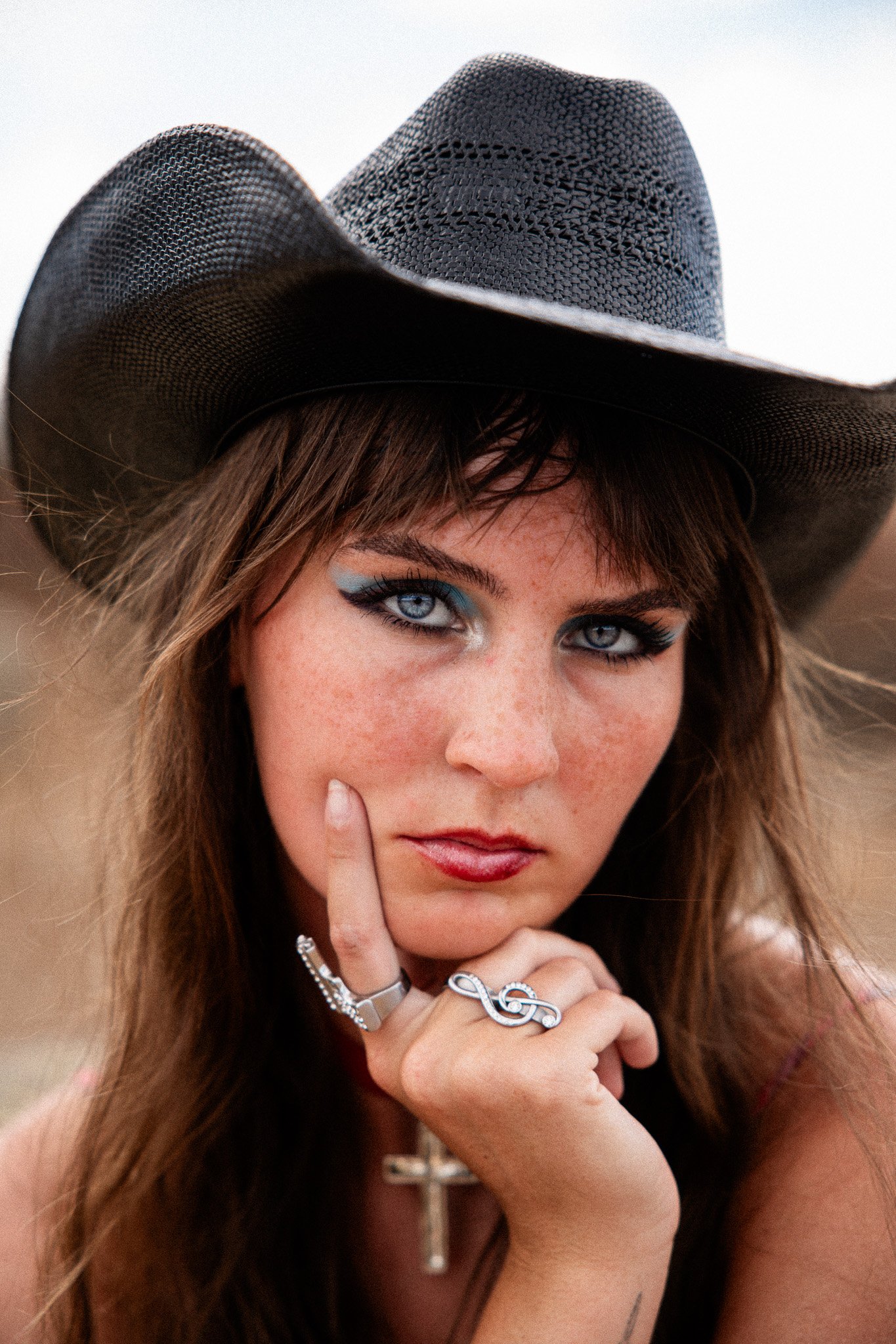 A woman with blue eye makeup, freckles, and long brown hair wearing a black wide-brimmed hat and jewelry, including rings and a cross necklace, resting her chin on her finger.