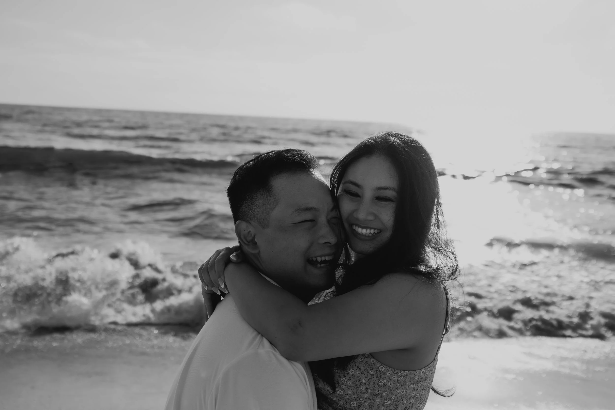 A black-and-white photo of a happy couple hugging on a beach, with ocean waves in the background.