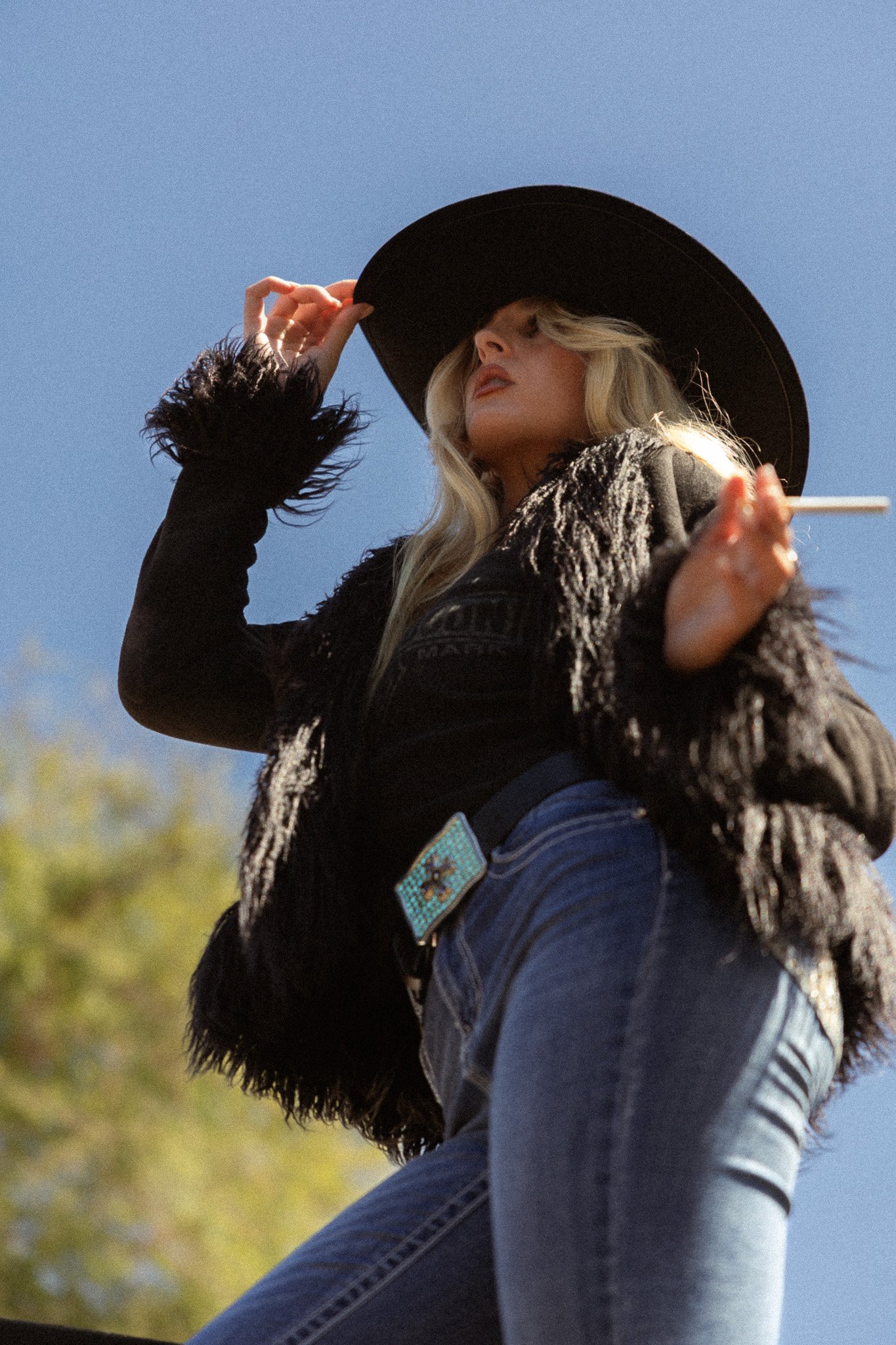 A woman wearing a large black hat, a fuzzy black jacket, and blue jeans standing outdoors against a blue sky and some trees, looking down and adjusting her hat.