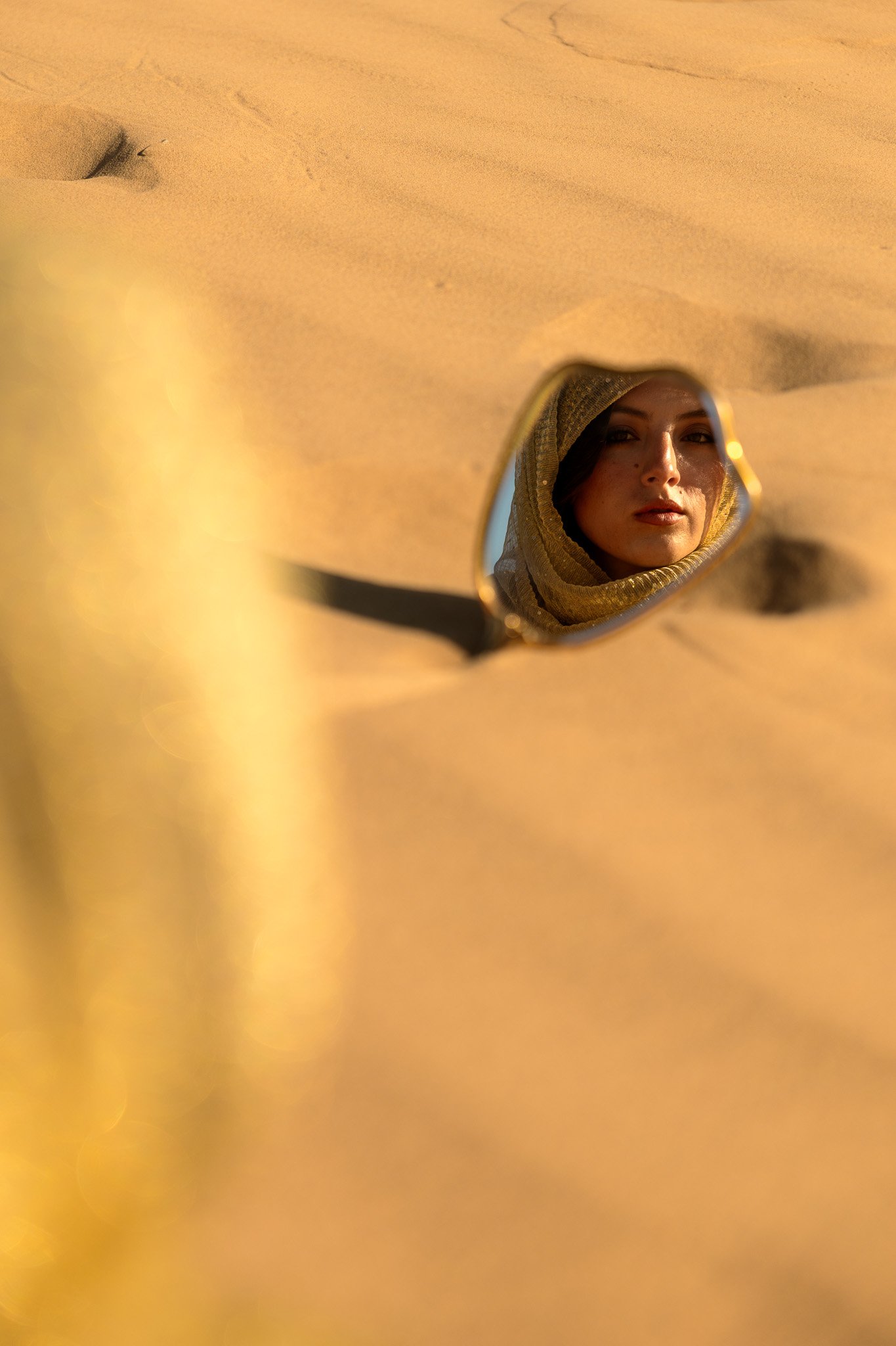A woman wearing a headscarf looking into a small, irregularly shaped mirror in a sandy area.