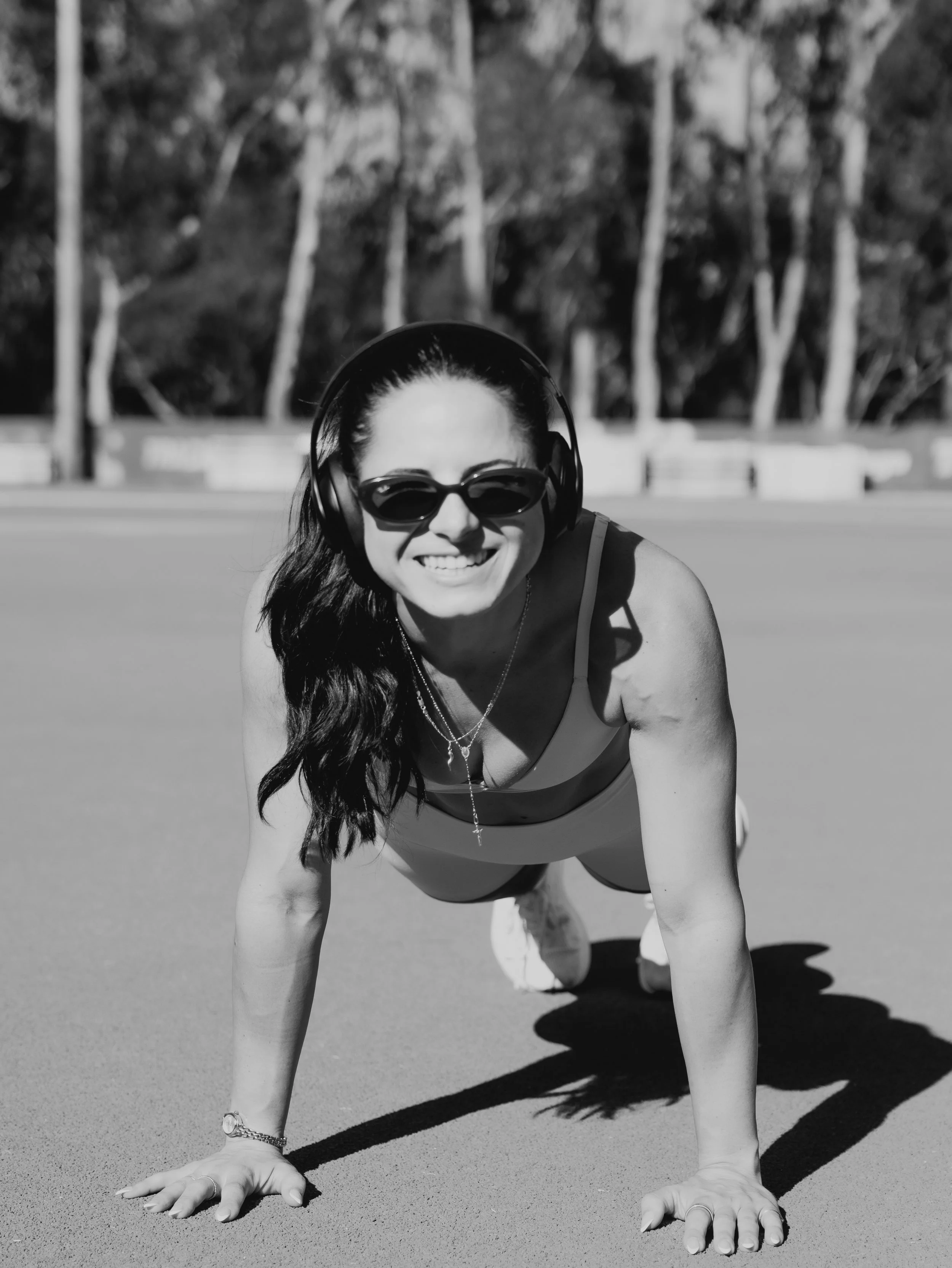 A woman with long hair, sunglasses, and headphones is doing a push-up on a sandy surface outdoors, with trees and a white fence in the background.