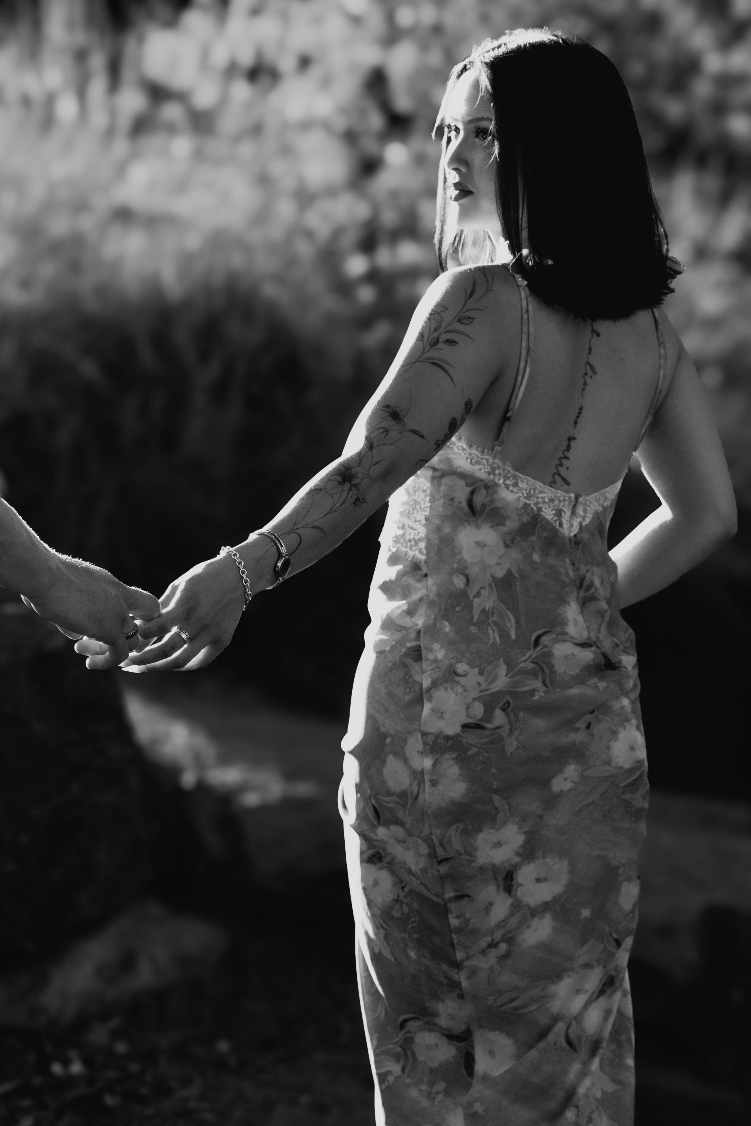 A black and white photo of a woman with dark hair, tattoos on her arm and back, dressed in a floral dress, holding hands with another person outdoors.